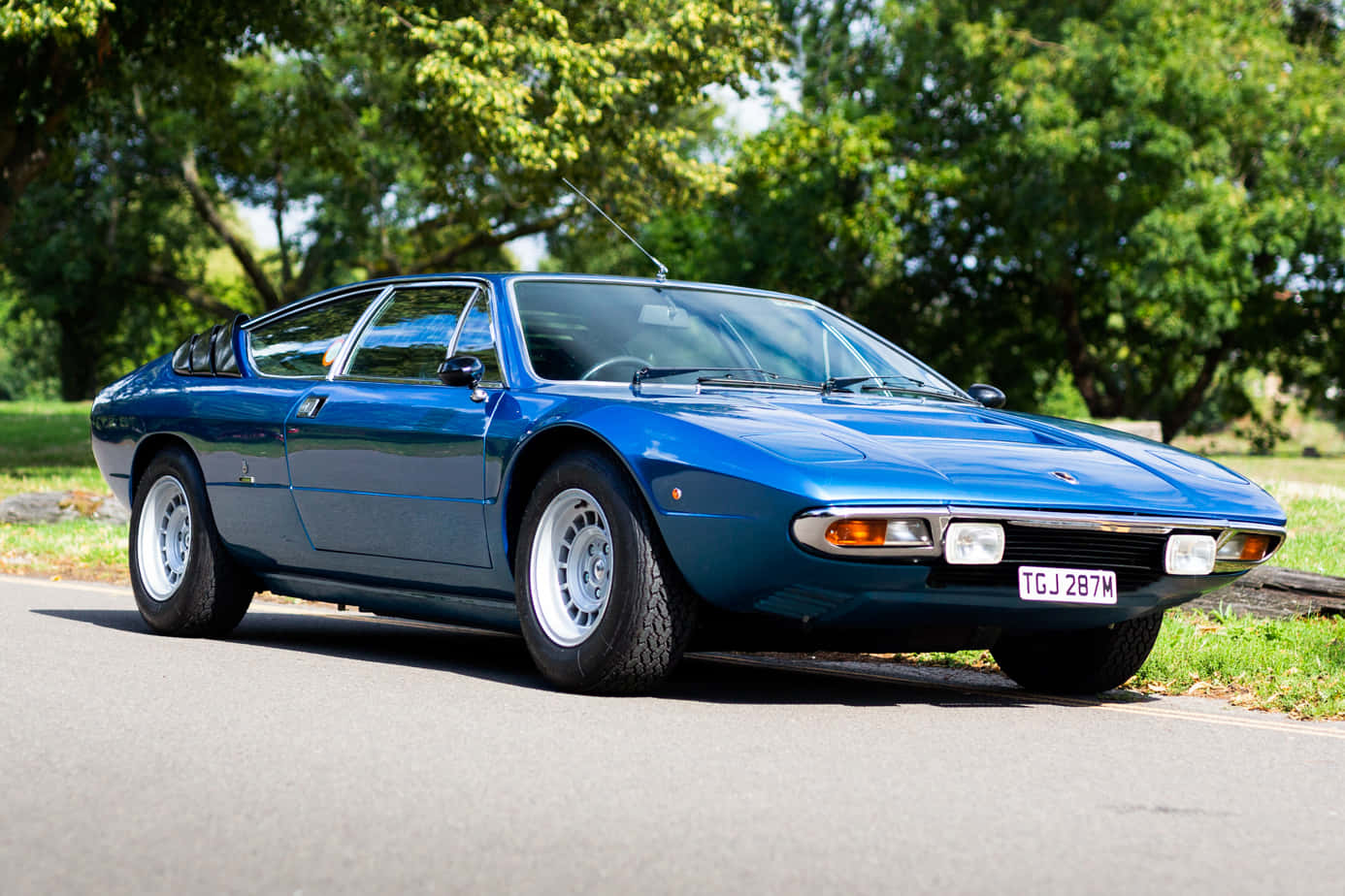 Lamborghini Urraco 桌布 壁紙 — 一架時尚的藍色經典跑車停泊在林蔭大道上，沐浴在明亮的日光下。
