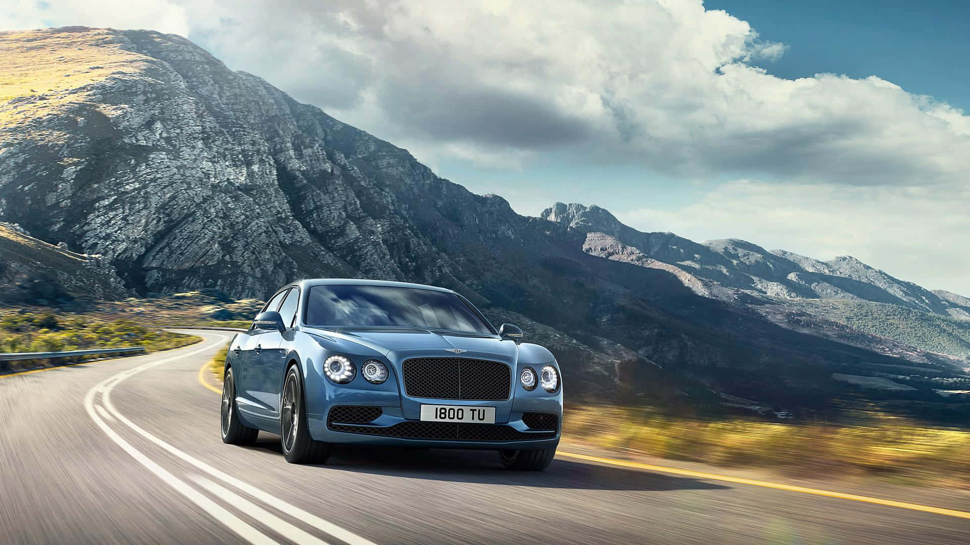 Bentley Flying Spur 桌布 壁紙 — 一架時尚的藍色賓利在戲劇性的多雲天空下沿著蜿蜒的山路疾馳。