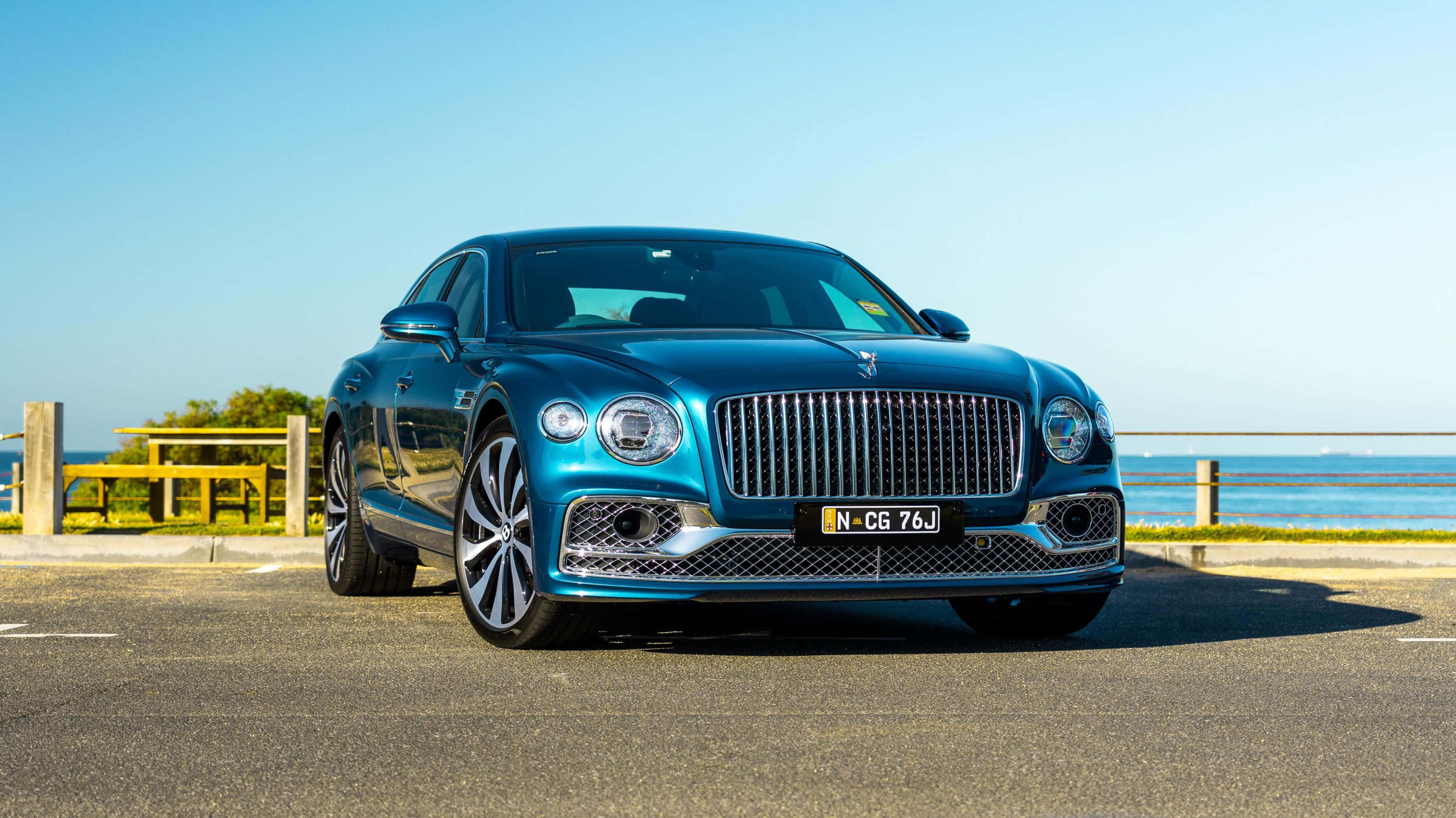 Bentley Flying Spur 桌布 壁紙 — 一架時尚的藍色賓利飛馳停泊在沿海道路上，背景是海洋和晴朗的天空。