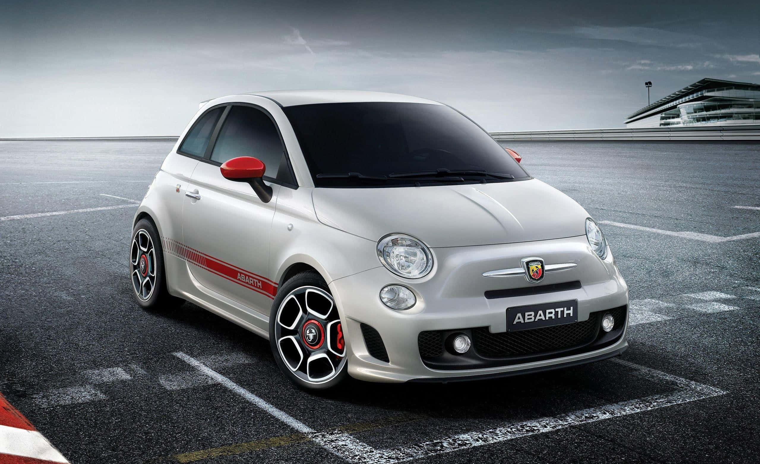 Abarth 桌布 壁紙 — 一架時尚的銀色 Abarth 跑車，帶有紅色點綴，停泊在陰天下的賽車場上。