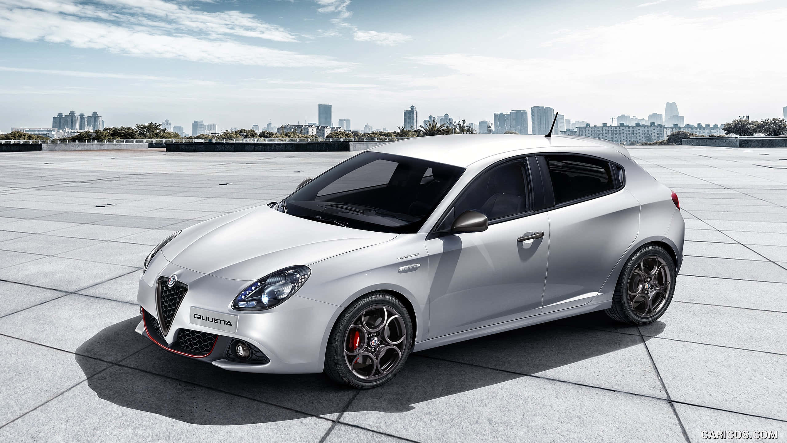 Alfa Romeo Giulietta 桌布 壁紙 — 一架時尚的銀色 Alfa Romeo Giulietta 跑車停泊在瓷磚屋頂上，背景是晴朗天空下的城市天際線。