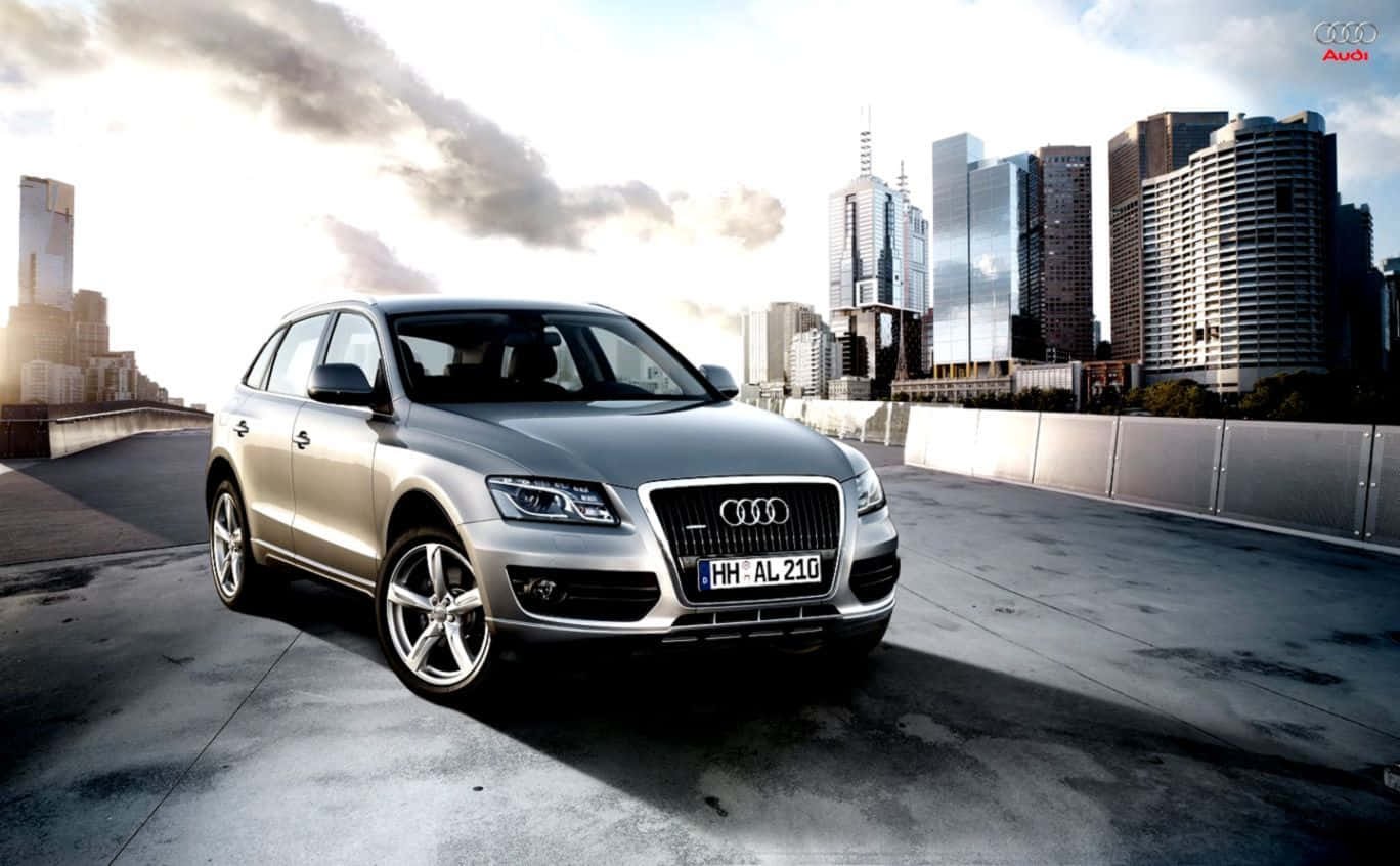 Audi Sq5 桌布 壁紙 — 一架時尚的銀色 Audi Q7 SUV 停泊在混凝土天橋上，背景是現代城市天際線和戲劇性的雲層。