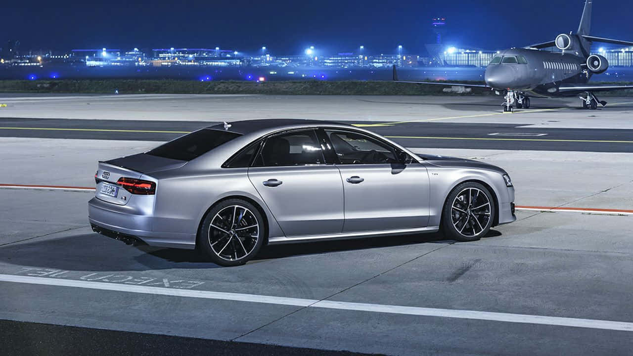Audi S8 桌布 壁紙 — 一架時尚的銀色 Audi 房車在夜間停在機場停機坪上，背景有私人飛機和城市燈光。