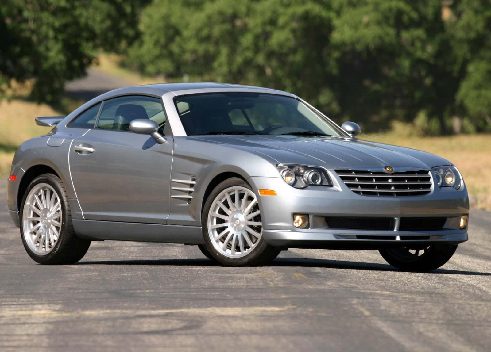 Chrysler Crossfire 桌布 壁紙 — 一架時尚的銀色 Chrysler Crossfire 跑車停在瀝青路上，背景有綠樹。