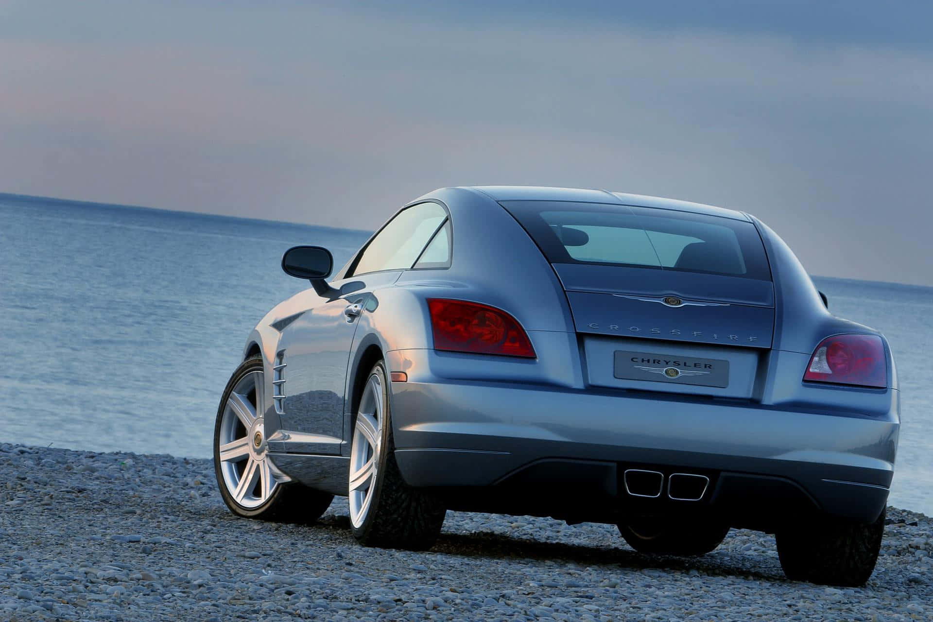 Chrysler Crossfire 桌布 壁紙 — 一架時尚的銀色 Chrysler Crossfire 跑車停泊在卵石海灘上，背景是海洋和暮色天空。