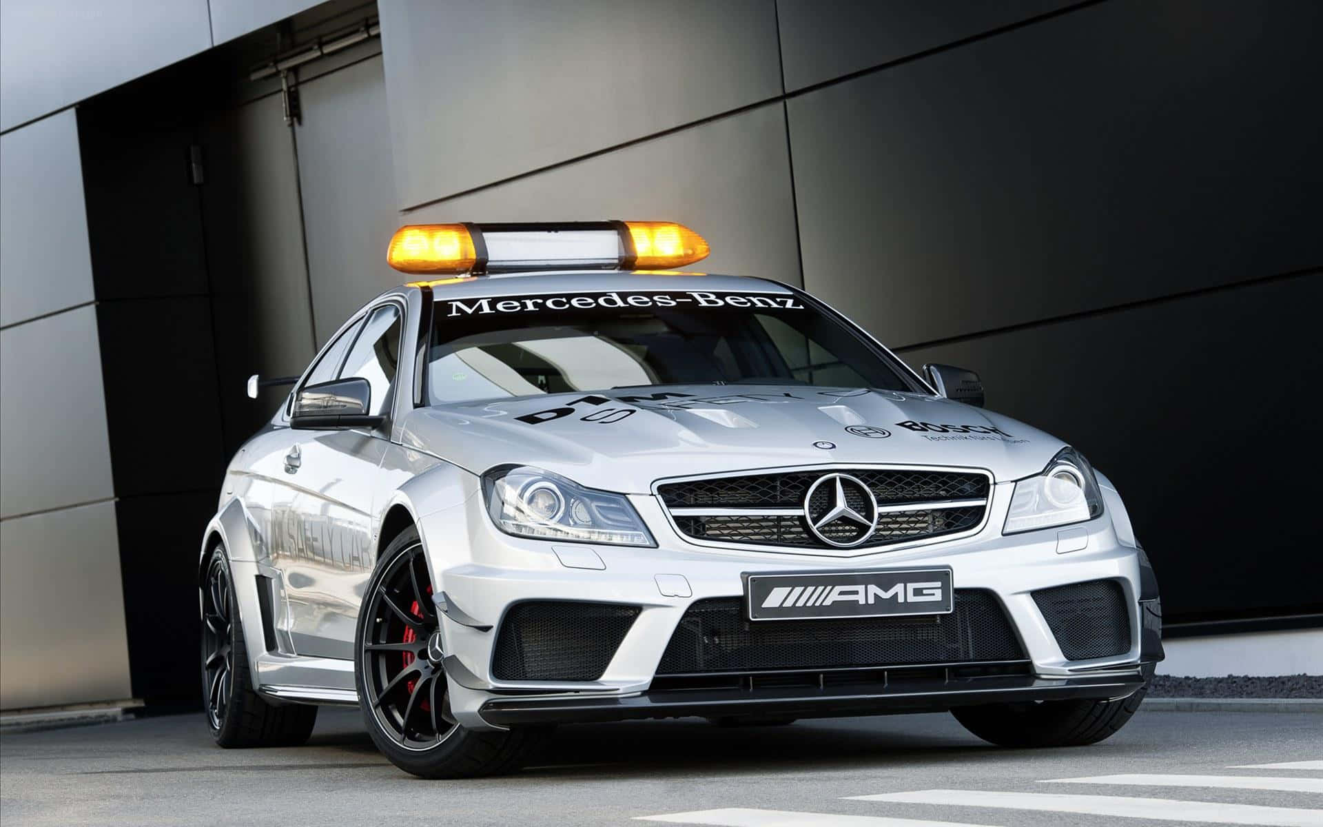 Safety Car 桌布 壁紙 — 一架時尚的銀色 Mercedes-AMG 賽車，帶有閃爍的琥珀色燈光，停在一棟現代建築前。