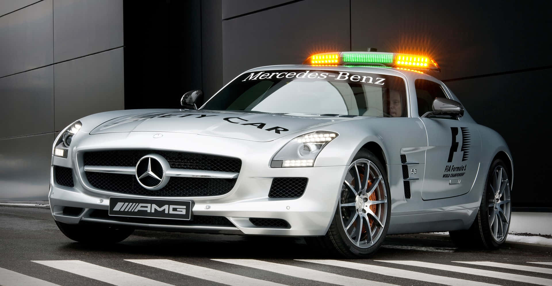 Safety Car 桌布 壁紙 — 一架時尚的銀色 Mercedes-AMG SLS GT1 安全車，閃爍著燈光，停泊在斑馬線上，背景是一座深色現代建築。