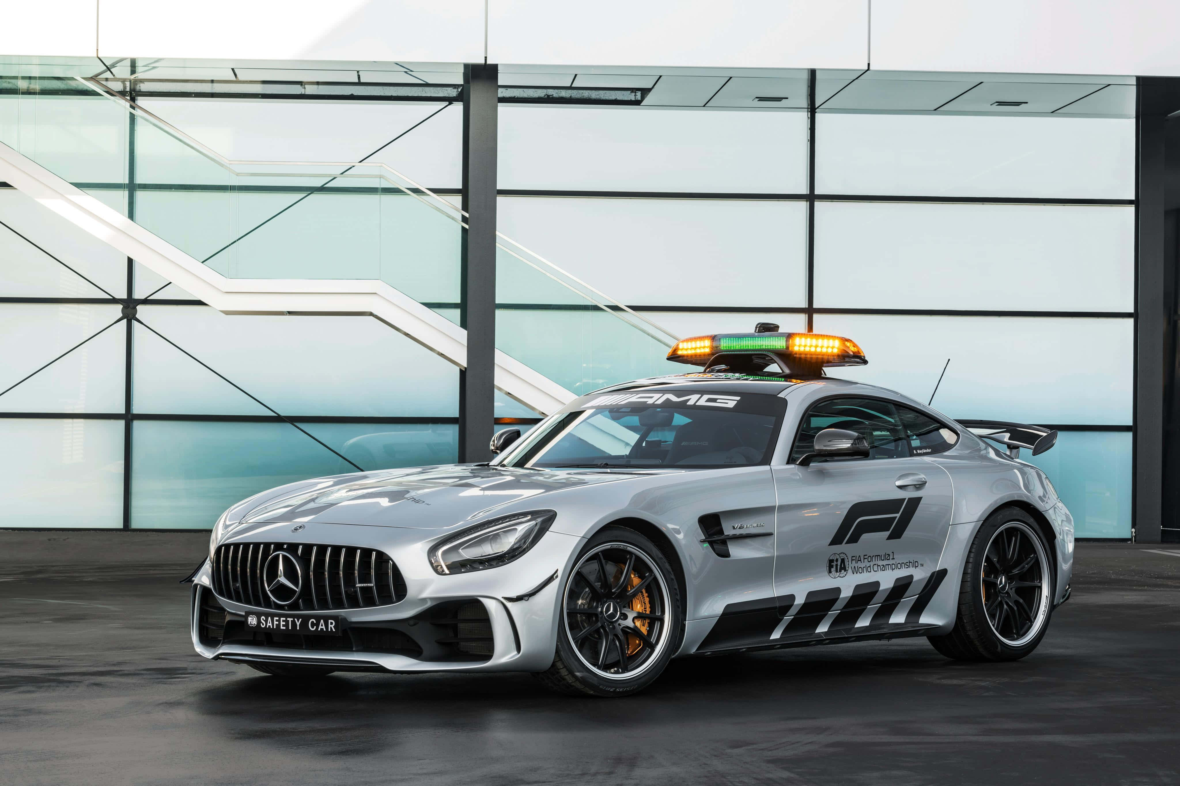 安全車帶領賽事 壁紙 — 一架時尚的銀色 Mercedes-AMG GT 安全車，閃爍著燈光，停在一棟現代玻璃建築前。