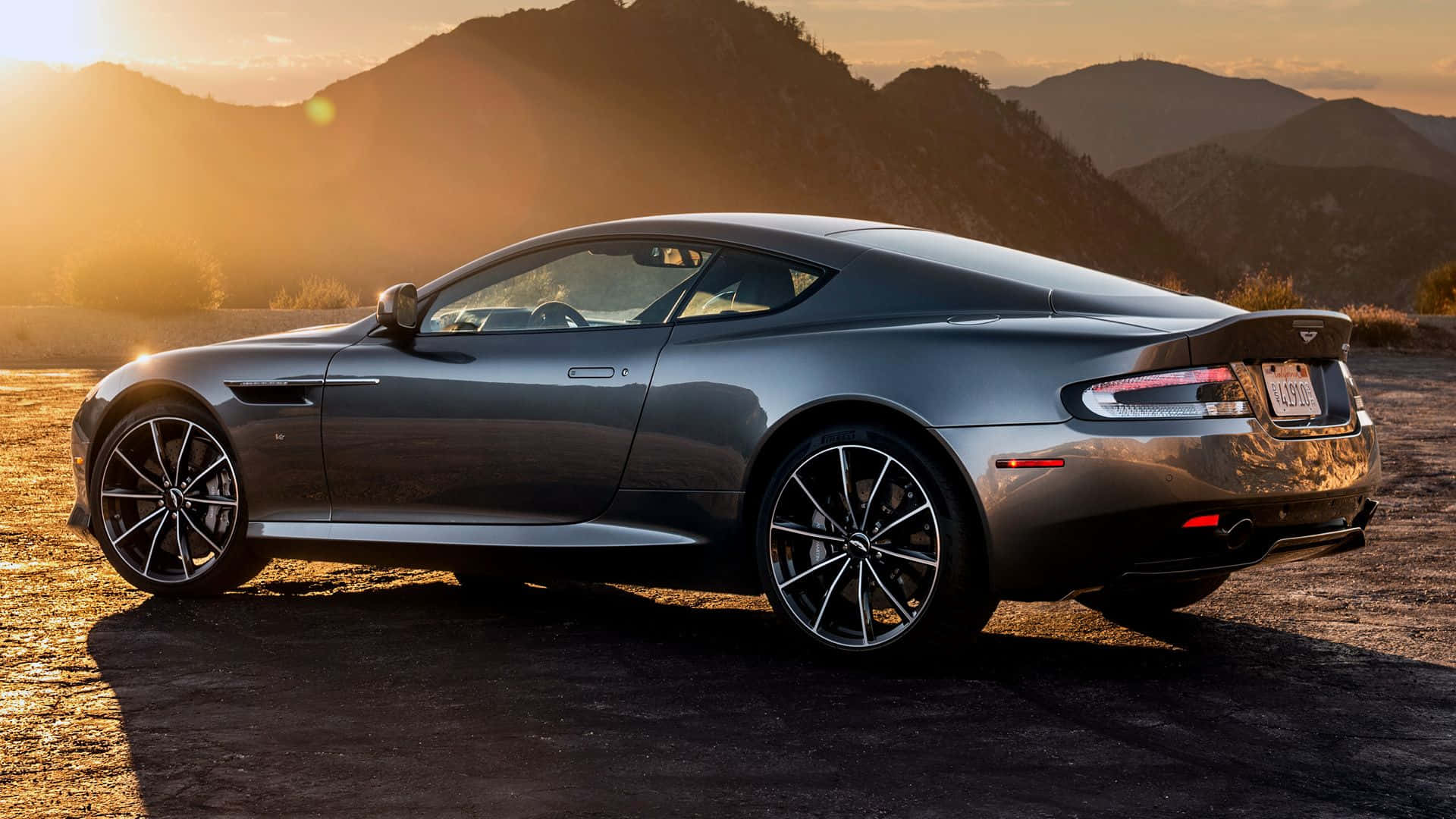 Aston Martin Db9 桌布 壁紙 — 一架時尚的銀色Aston Martin跑車停泊在塵土飛揚的道路上，群山沐浴在金色的日落光芒中。