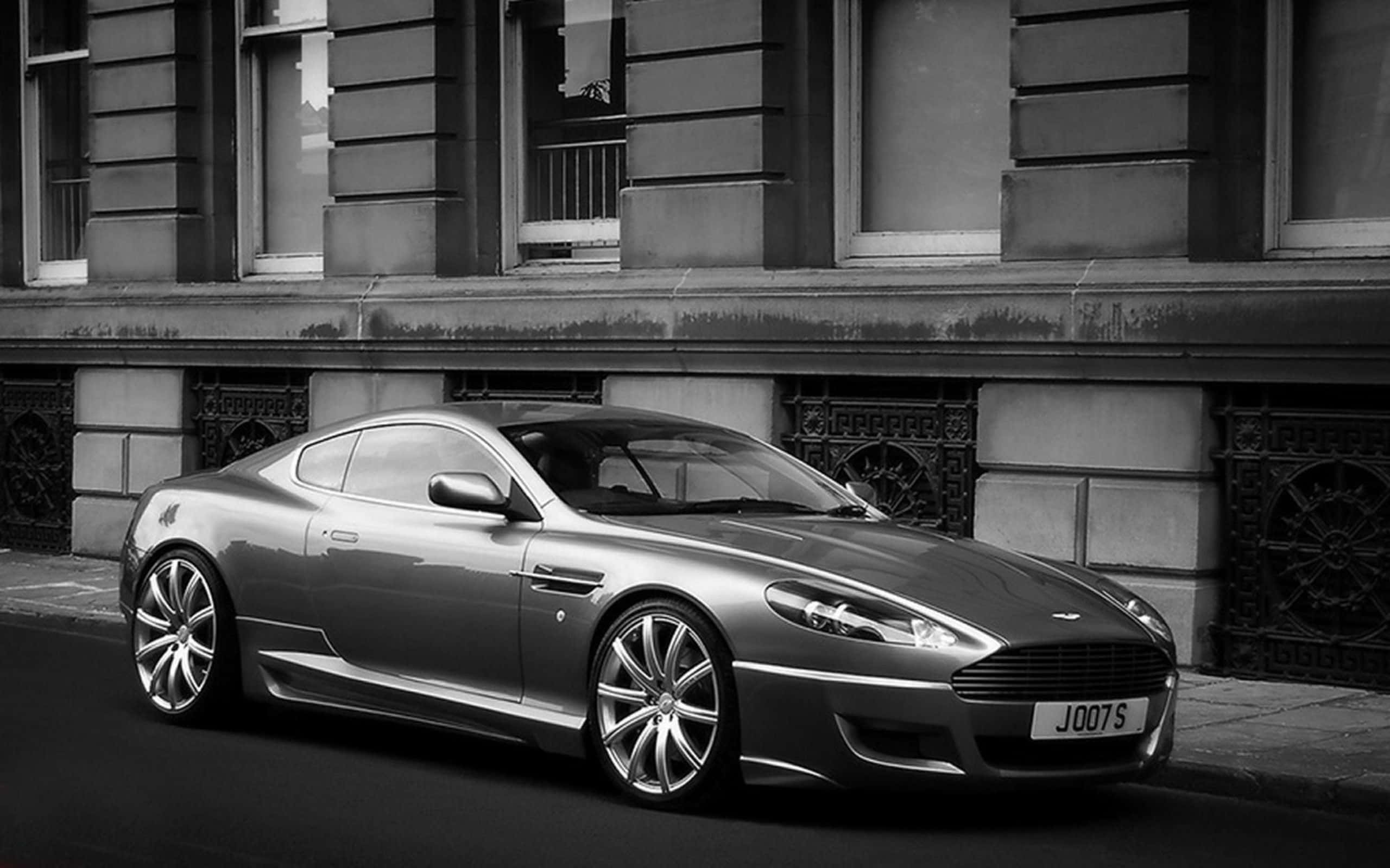 Aston Martin Db9 桌布 壁紙 — 一架時尚的銀色Aston Martin DB9停泊在城市街道上，前面是經典的石建築，黑白風格。