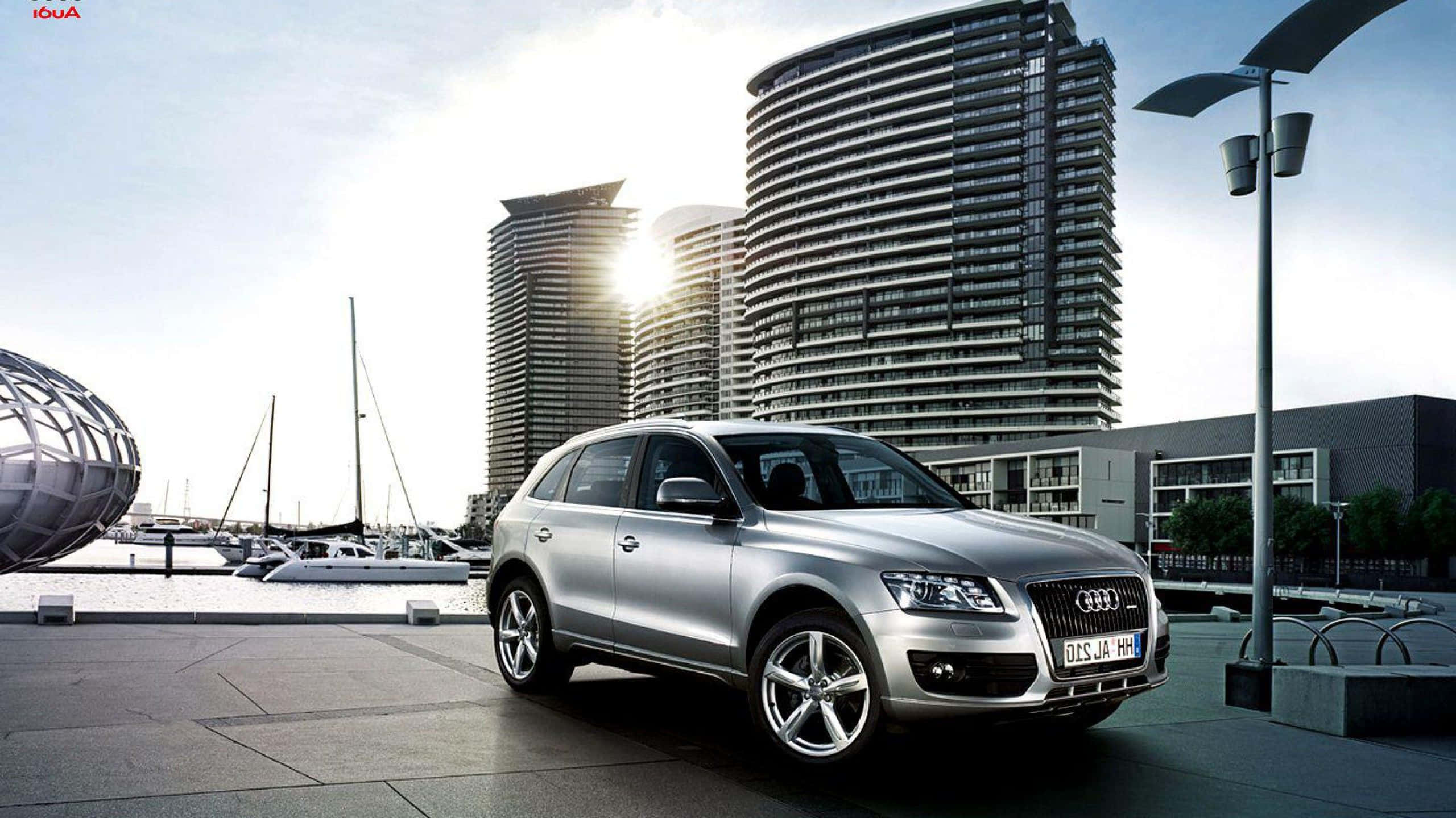 Audi Sq5 桌布 壁紙 — 一架時尚的銀色Audi Q7 SUV停泊在現代海濱廣場上，背景有彎曲的玻璃塔和帆船。
