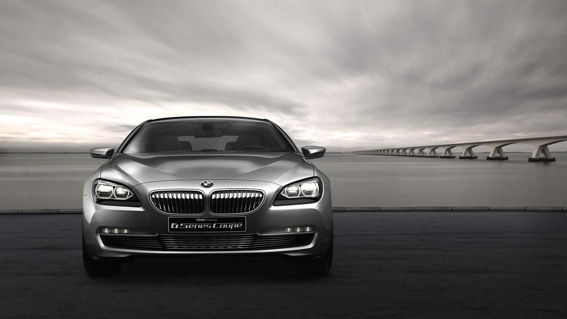 Bmw 6 Series 桌布 壁紙 — 一架時尚的銀色BMW 6系轎跑車在黑暗的道路上向前行駛，一條長橋延伸至水面上多雲的天空。