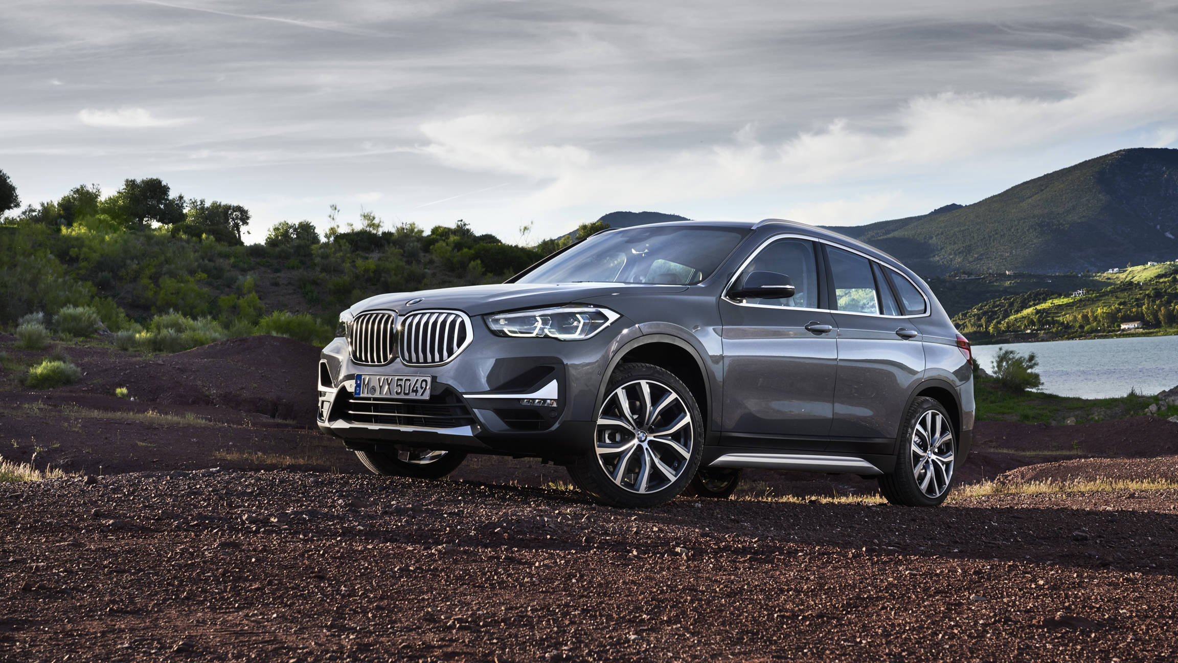 road trip 桌布 壁紙 — 一架時尚的銀色BMW X1 SUV停泊在碎石地形上，旁邊有湖泊和綠色山丘，天空多雲。