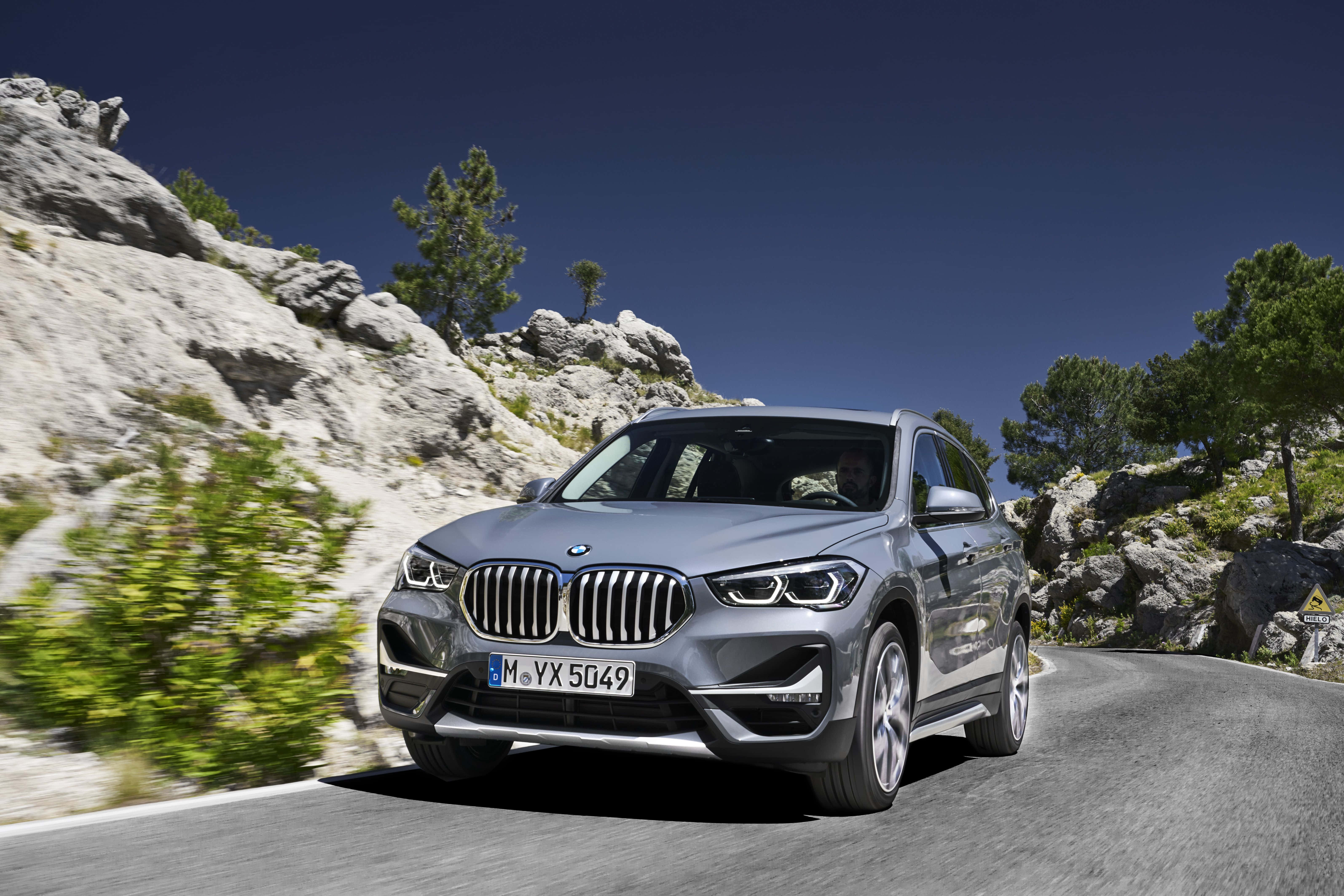 Bmw I8 桌布 壁紙 — 一架時尚的銀色BMW X1 SUV在晴朗的藍天下，沿著蜿蜒的山路行駛，兩旁是岩石峭壁和綠樹。