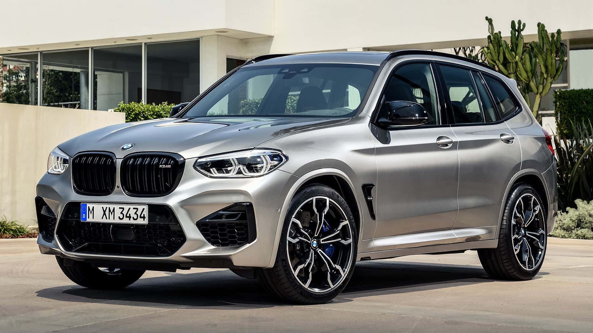Bmw X3 桌布 壁紙 — 一架時尚的銀色BMW X3 M停泊在陽光普照的車道上，旁邊是一座有仙人掌和綠植的現代房屋。