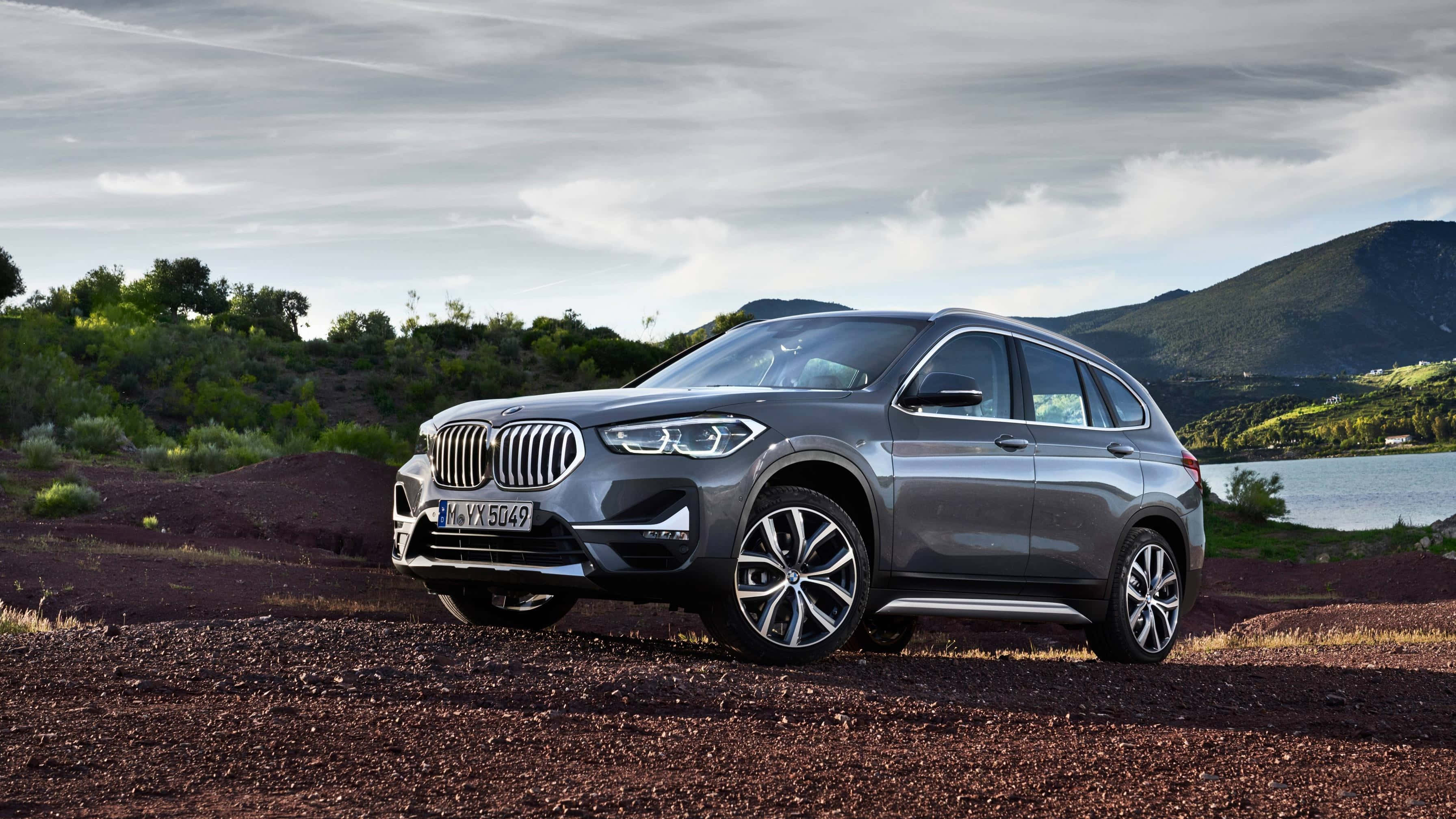 Bmw X1 桌布 壁紙 — 一架時尚的銀色BMW X3 SUV停泊在平靜湖泊旁的紅褐色泥路上，背景是綠丘和陰天。