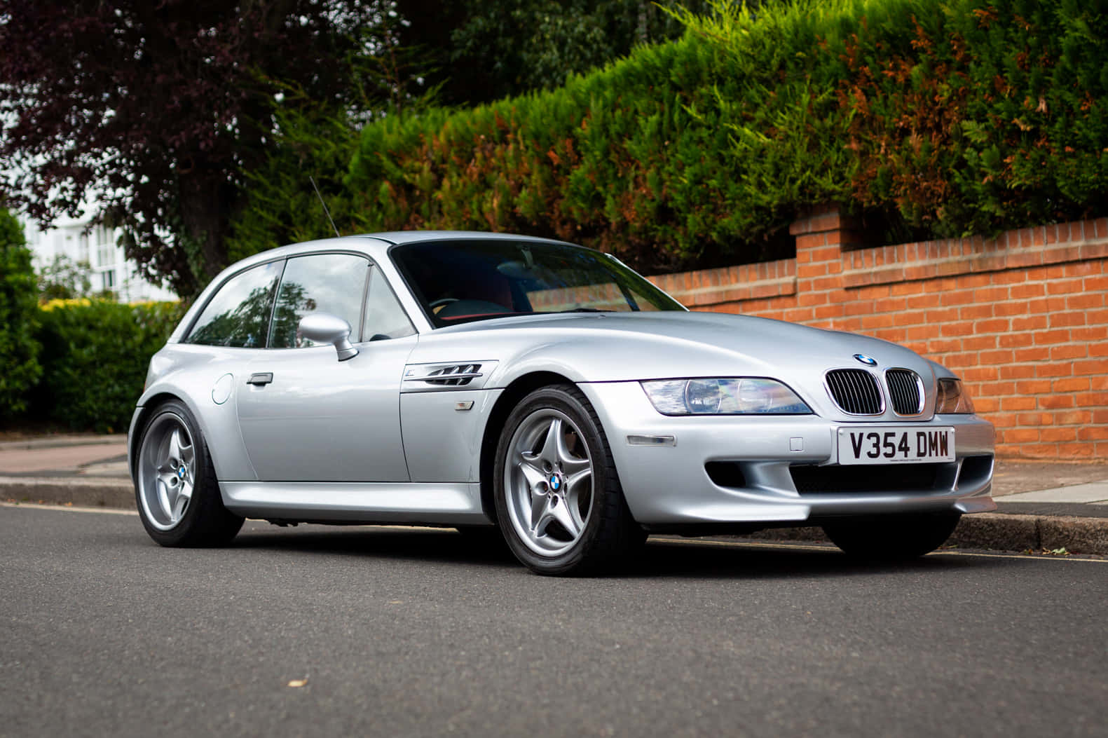 Bmw Z3 桌布 壁紙 — 一架時尚的銀色BMW Z3 M Coupe停泊在寧靜的住宅街道上，旁邊是磚牆和綠色籬笆。