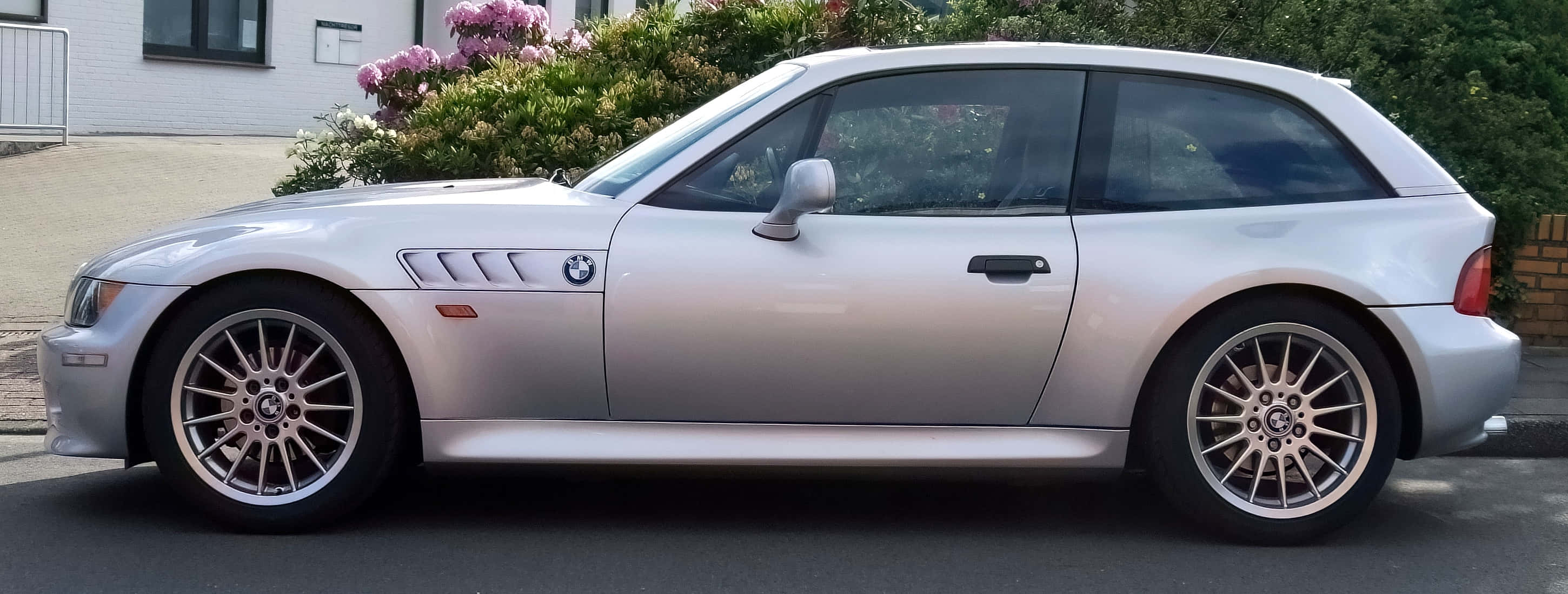 Bmw Z3 桌布 壁紙 — 一架時尚的銀色BMW Z3開篷跑車停泊在住宅街道上，背景有盛開的粉紅色花朵和綠色灌木。