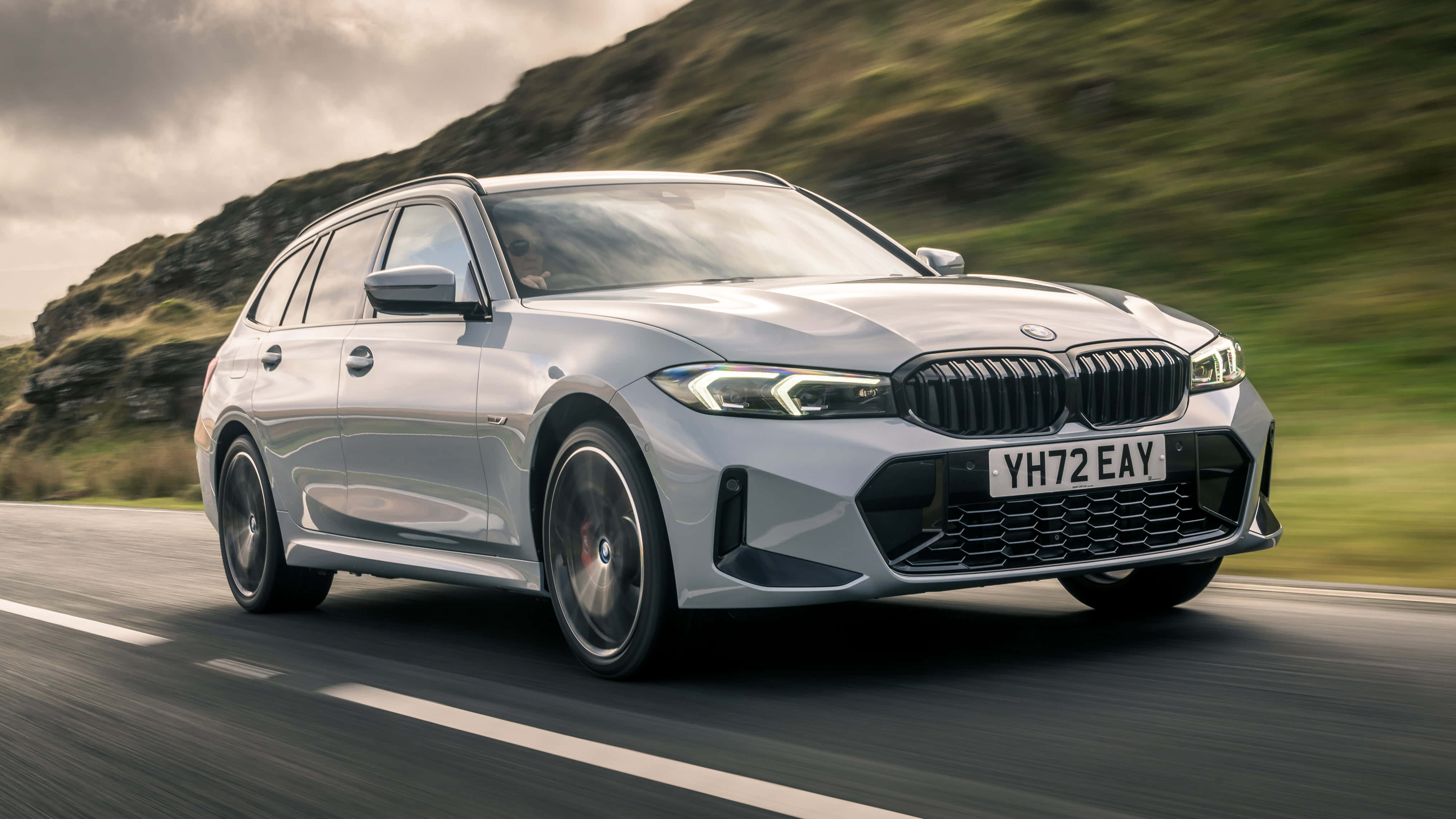 Bmw 3 Series 桌布 壁紙 — 一架時尚的銀色BMW旅行車沿著蜿蜒的道路疾馳，背景是模糊的綠色山丘。