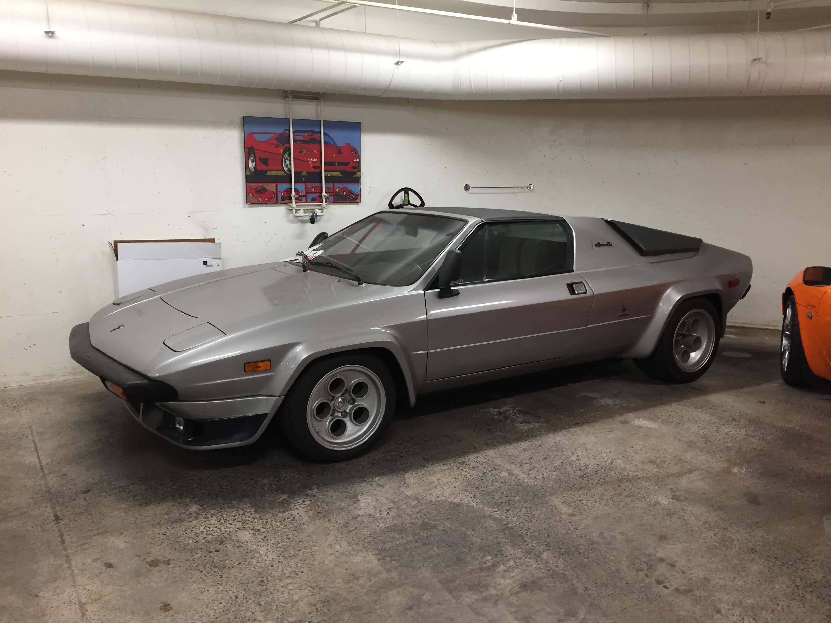 Lamborghini Silhouette 桌布 壁紙 — 一架時尚的銀色DeLorean跑車停泊在混凝土車庫中，車庫有外露管道和牆上的紅色汽車海報。