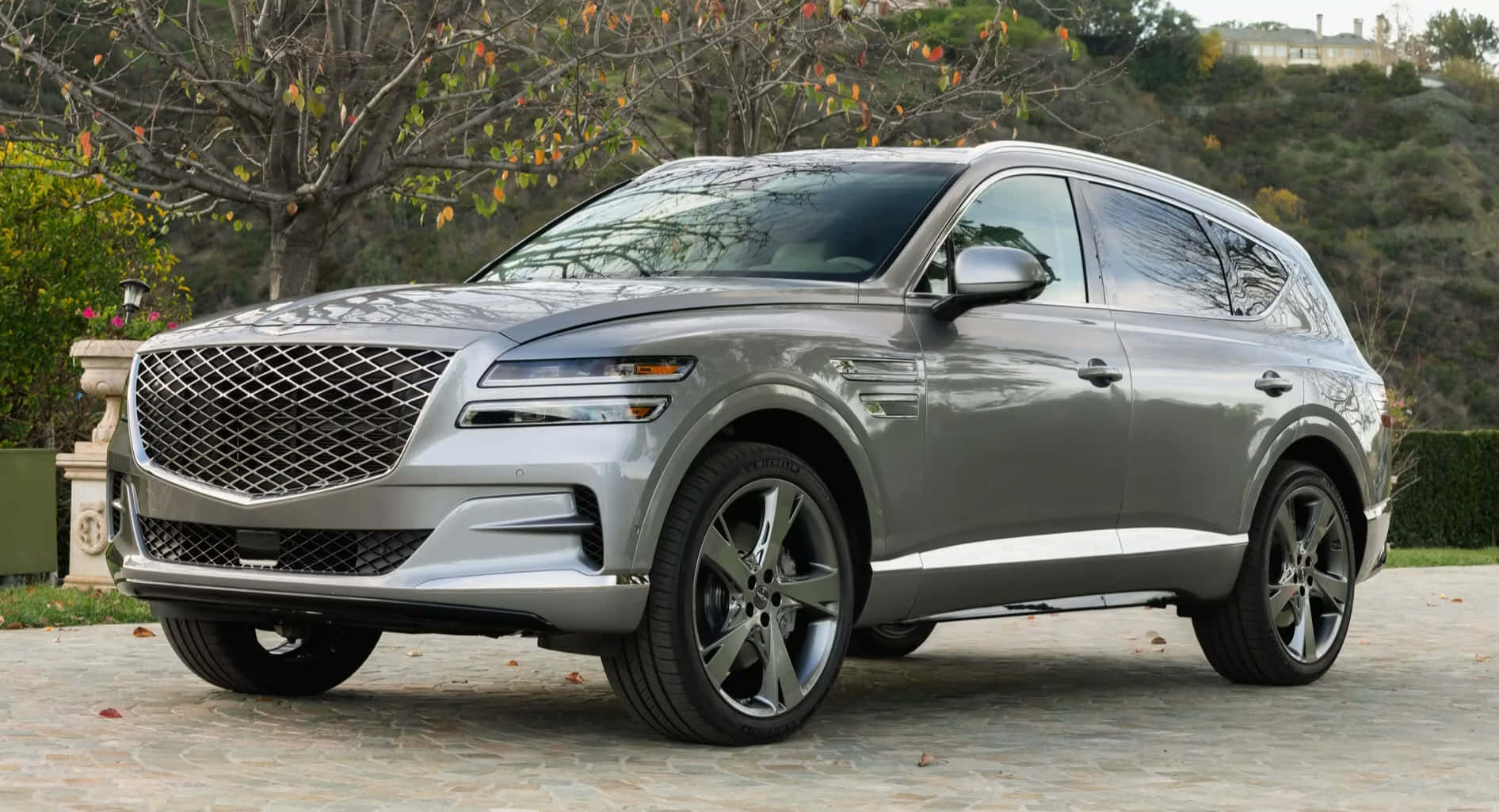 Genesis Gv80 桌布 壁紙 — 一架時尚的銀色Genesis GV80 SUV停泊在石頭車道上，背景有秋天的樹木和山丘。