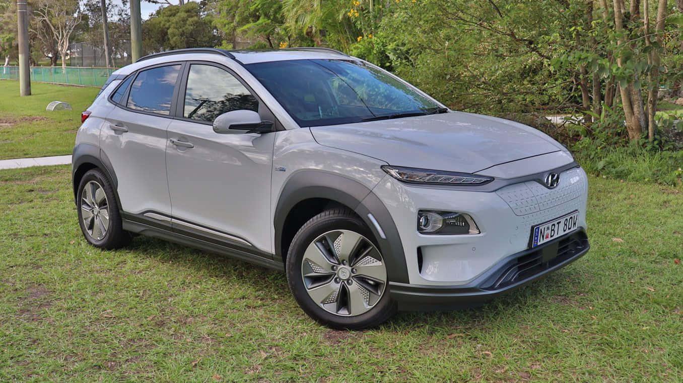 Electric Vehicle 桌布 壁紙 — 一架時尚的銀色Hyundai Kona Electric SUV停泊在草地上，背景有樹木和圍欄。