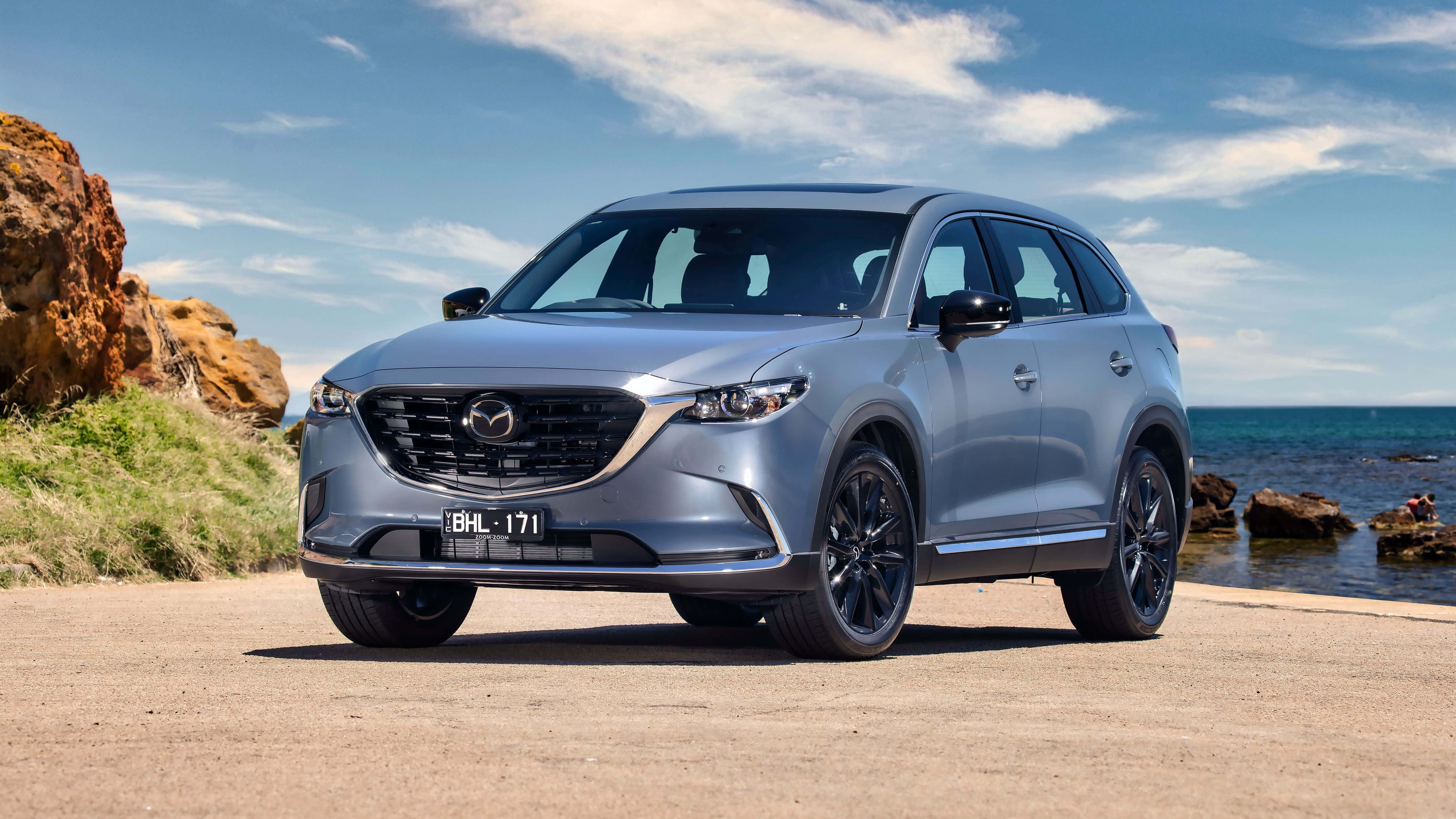 Mazda Cx-9 桌布 壁紙 — 一架時尚的銀色Mazda CX-90 SUV停泊在沿海道路上，背景是岩石峭壁和海洋，上方是明亮的藍天。