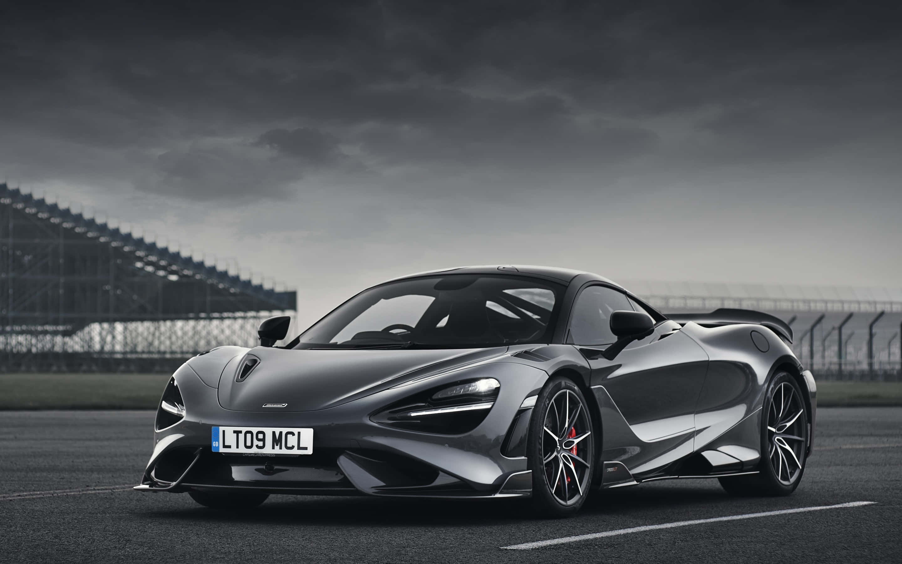 麥拿侖 桌布 壁紙 — 一架時尚的銀色McLaren跑車停在賽道上，背景是陰沉多雲的天空和看台。