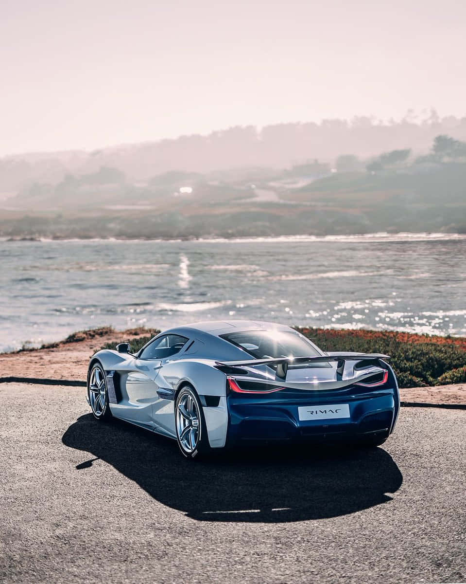 Rimac 桌布 壁紙 — 一架時尚的銀色Rimac超跑停泊在沿海道路上，背景有海浪和朦朧的山丘。