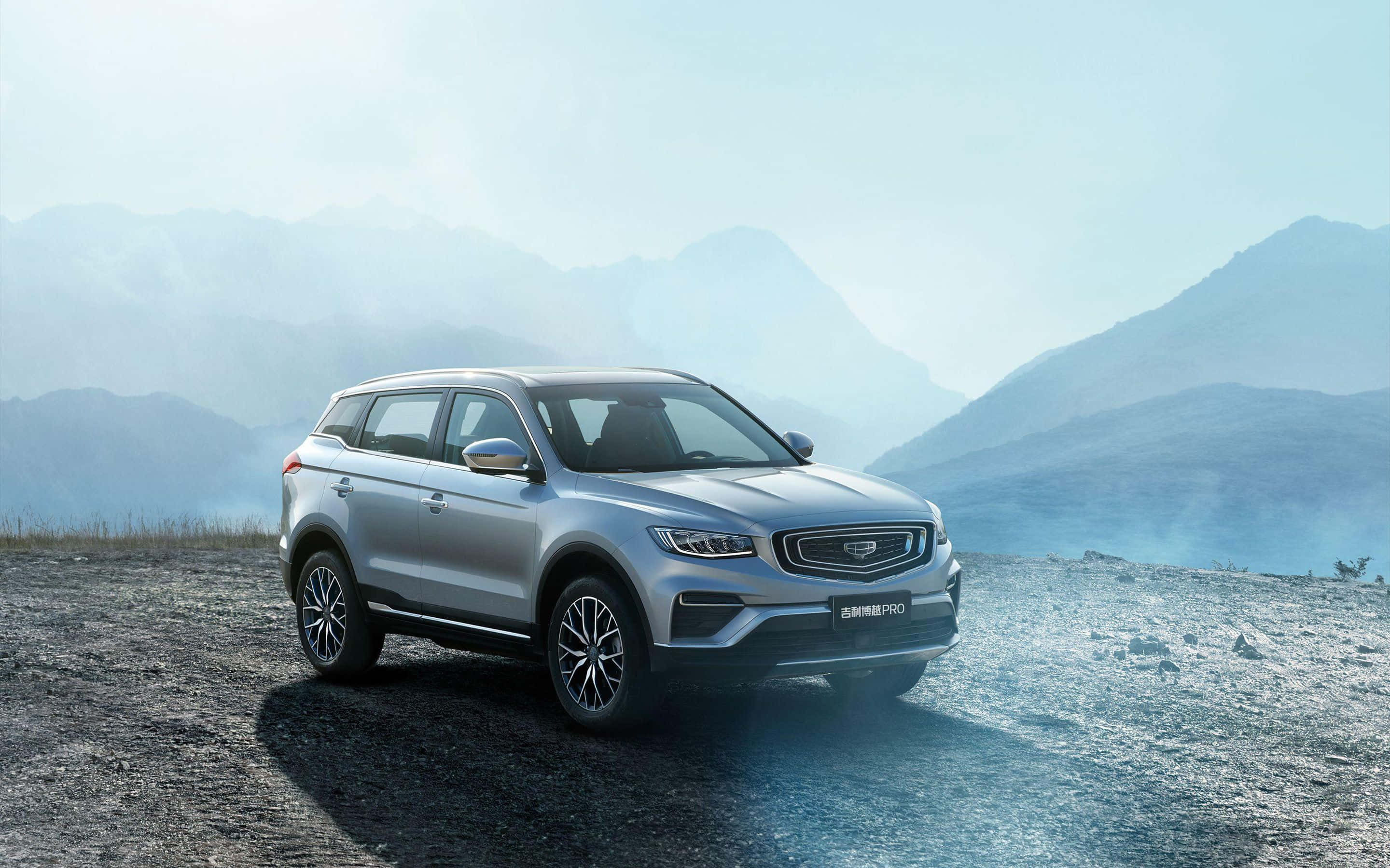 Geely 桌布 壁紙 — 一架時尚的銀色SUV停泊在崎嶇的山景瞭望點，背景是霧氣繚繞的山峰，天空柔和湛藍。