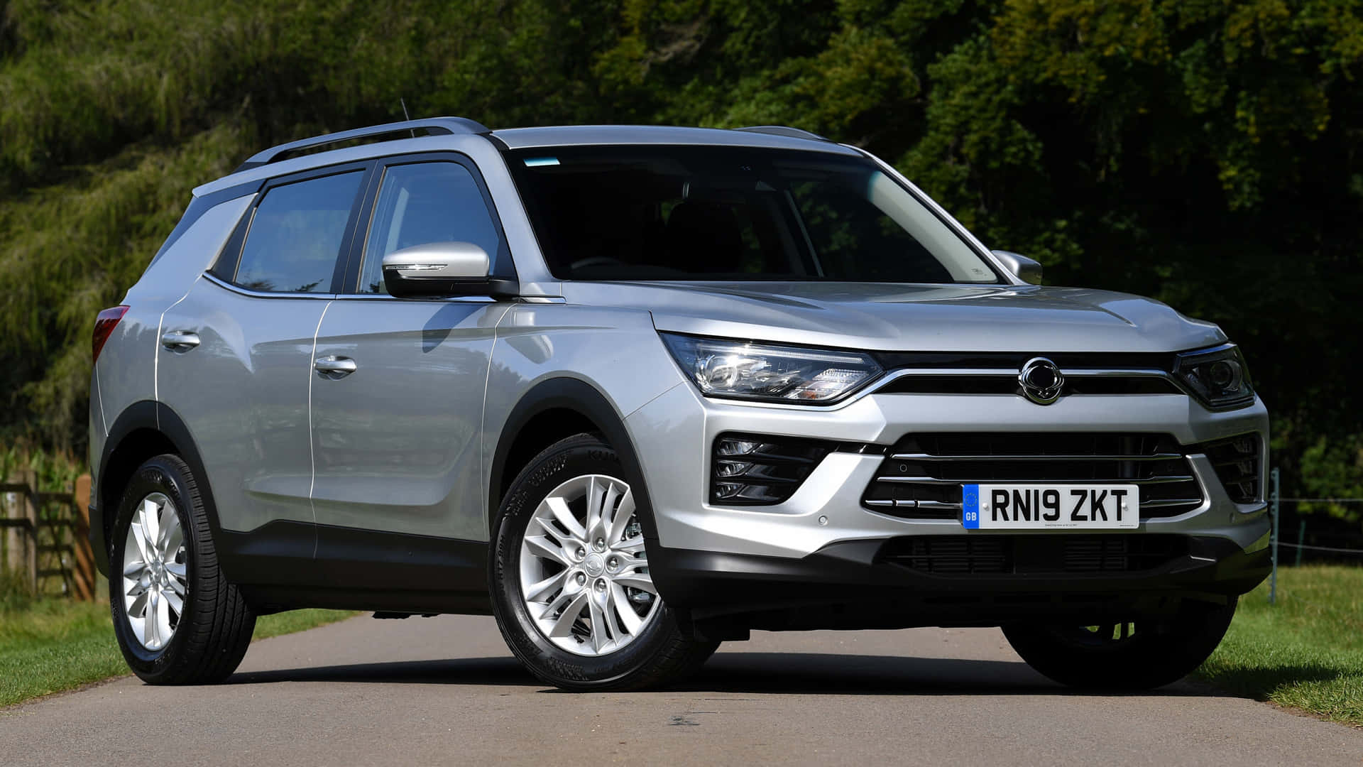 Ssangyong 桌布 壁紙 — 一架時尚的銀色SUV停泊在鋪砌的道路上，背景有茂密的綠樹和草地，在明亮的日光下。
