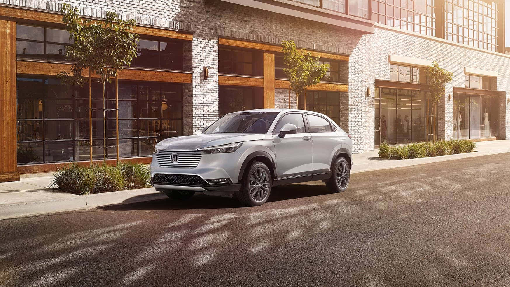 Honda Hr-v 桌布 壁紙 — 一架時尚的銀色SUV停泊在陽光普照的城市街道上，旁邊是帶有大型玻璃窗的現代磚砌建築。