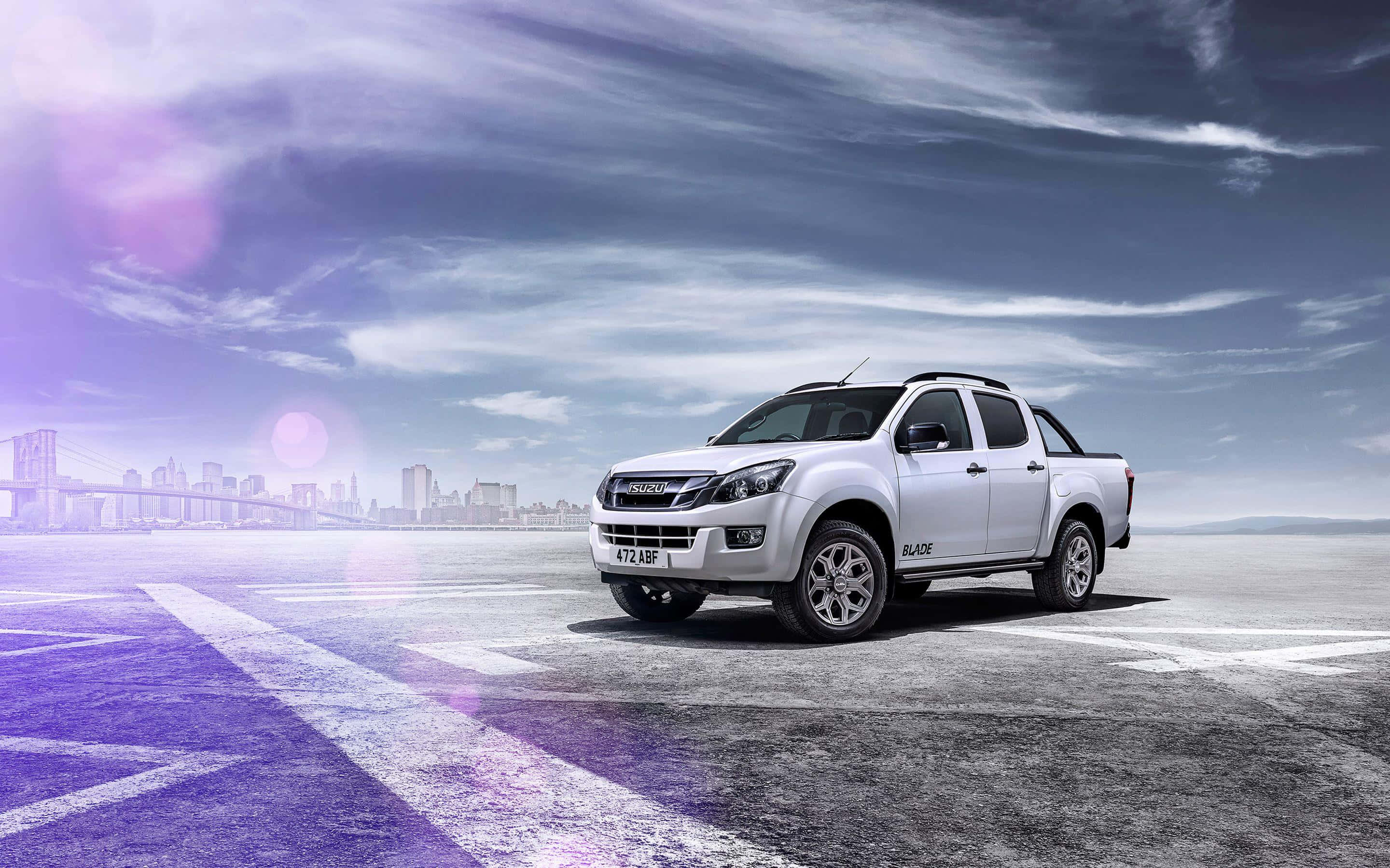 Isuzu 桌布 壁紙 — 一架時尚的銀色五十鈴皮卡停泊在廣闊的鋪砌區域，背景是城市天際線和橋樑，天空呈現戲劇性的紫色和藍色。