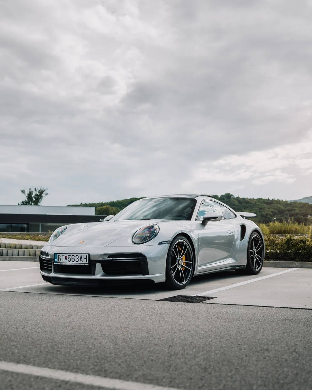 Sports Car 桌布 壁紙 — 一架時尚的銀色保時捷911 Turbo S停泊在空曠的停車場，天空多雲，背景是綠色山丘。