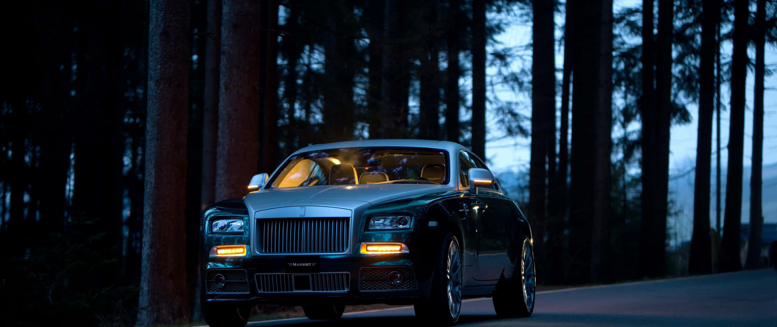 Rolls Royce 桌布 壁紙 — 一架時尚的銀色勞斯萊斯 Ghost 在暮色中停泊在森林道路上，車內溫暖的燈光閃爍。