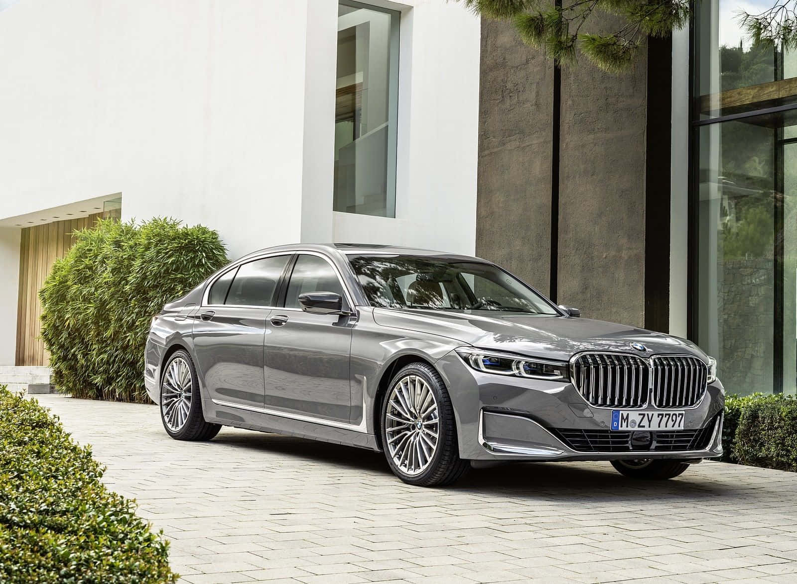 Bmw 7 Series 桌布 壁紙 — 一架時尚的銀色寶馬7系轎車停泊在石頭車道上，旁邊是一座現代白色和混凝土房屋，周圍有綠色灌木。