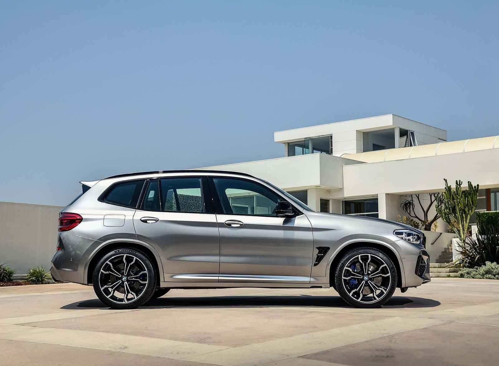 Bmw X3 桌布 壁紙 — 一架時尚的銀色寶馬X5 M停泊在陽光明媚的混凝土車道上，前方是帶有沙漠植物的現代白色房屋。