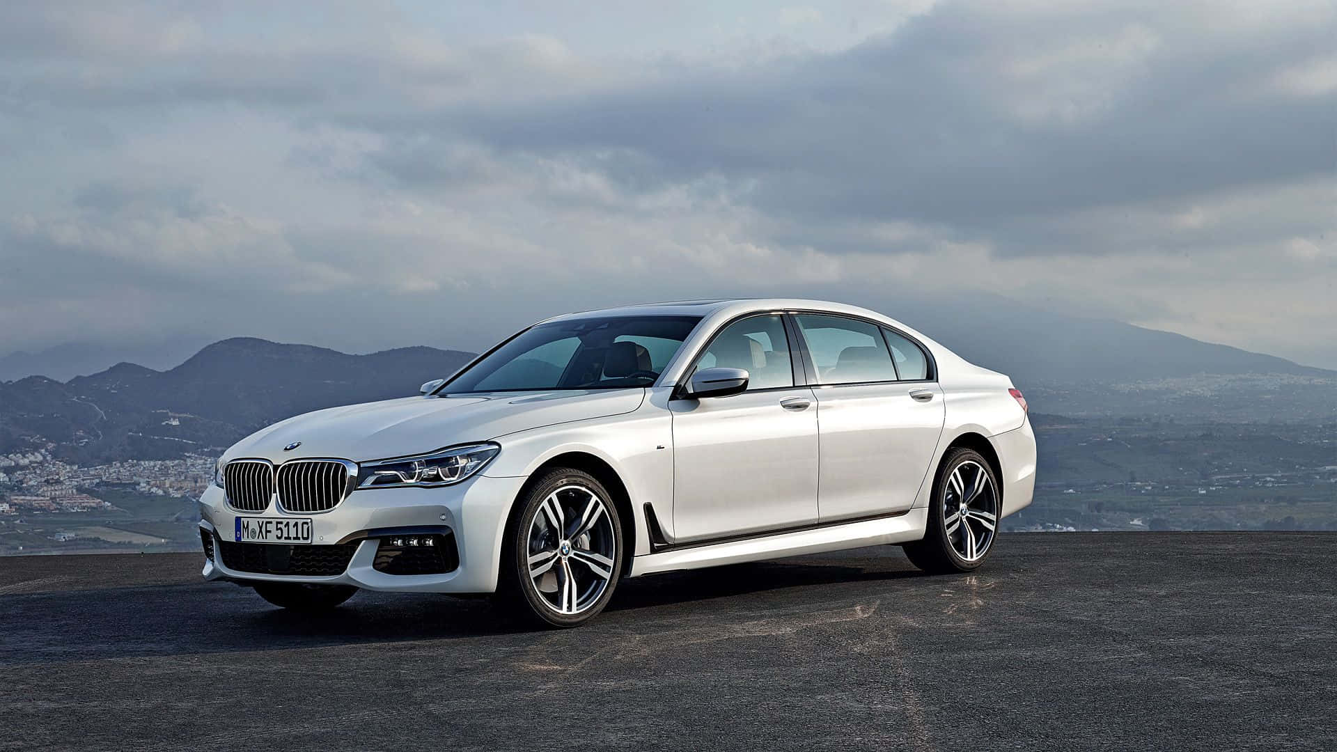 Bmw 7 Series 桌布 壁紙 — 一架時尚的銀色寶馬房車停泊在鋪砌的觀景點上，背景是遠山和陰天。