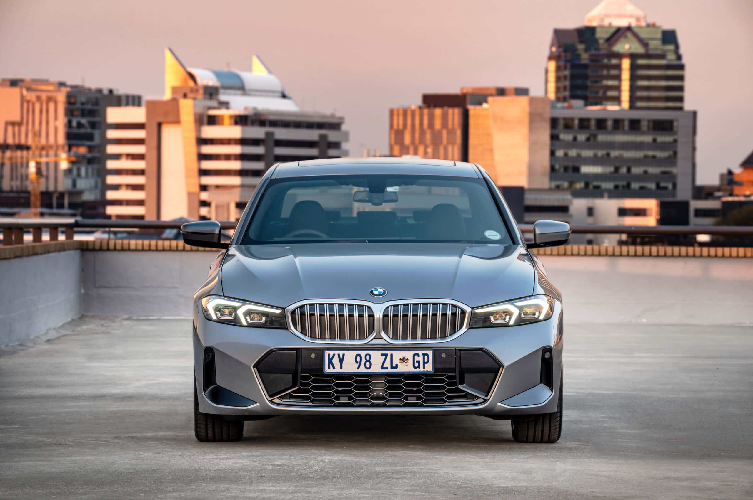Bmw 3 Series 桌布 壁紙 — 一架時尚的銀色寶馬房車在屋頂上向前方，背後是溫暖夕陽光輝映照的城市天際線。