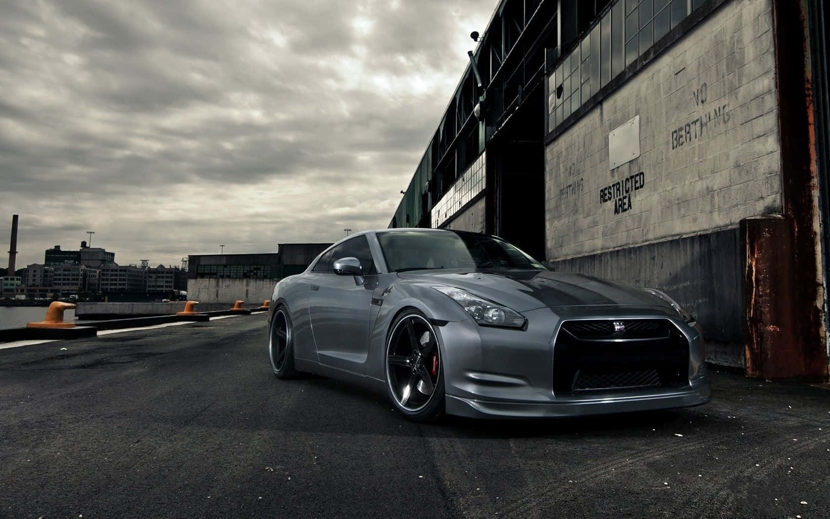 JDM 車 桌布 壁紙 — 一架時尚的銀色日產GT-R停泊在一個粗糙的碼頭上，旁邊是一座飽經風霜的工業建築，天空多雲。