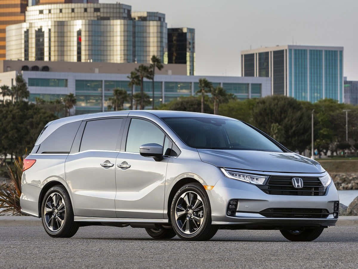 Honda Odyssey 桌布 壁紙 — 一架時尚的銀色本田小型貨車停在瀝青路上，背景有現代玻璃建築和棕櫚樹，在柔和日光下。