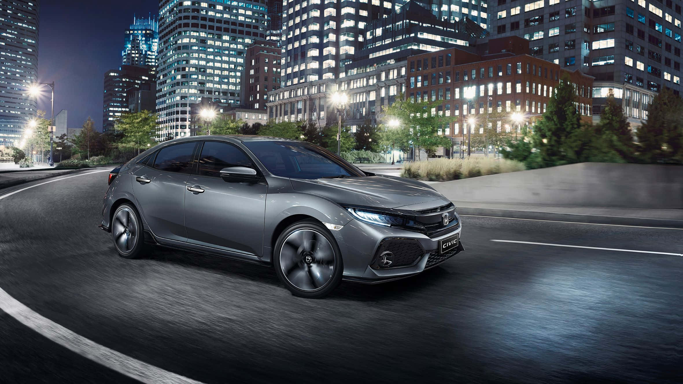 Hatchback 桌布 壁紙 — 一架時尚的銀色本田思域S在夜晚行駛於燈火通明的城市街道，周圍是高聳的摩天大樓。