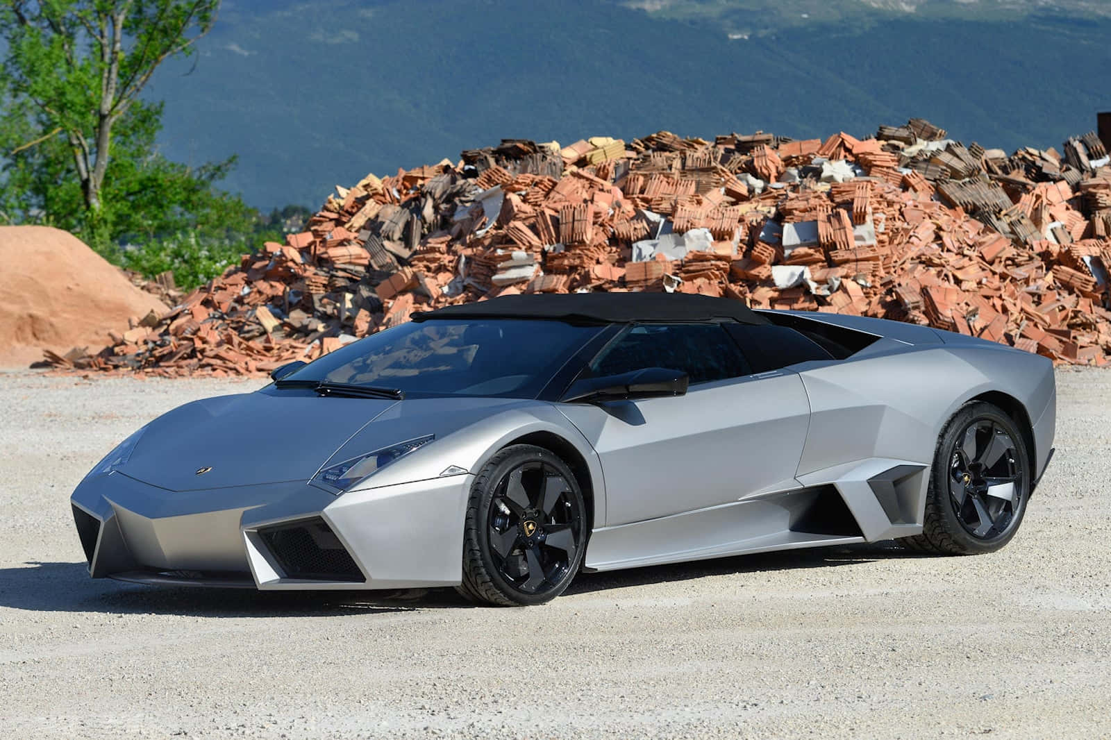 Lamborghini Reventón 桌布 壁紙 — 一架時尚的銀色林寶堅尼Reventon跑車停泊在碎石上，背景有一堆破碎的赤陶磚和綠樹。