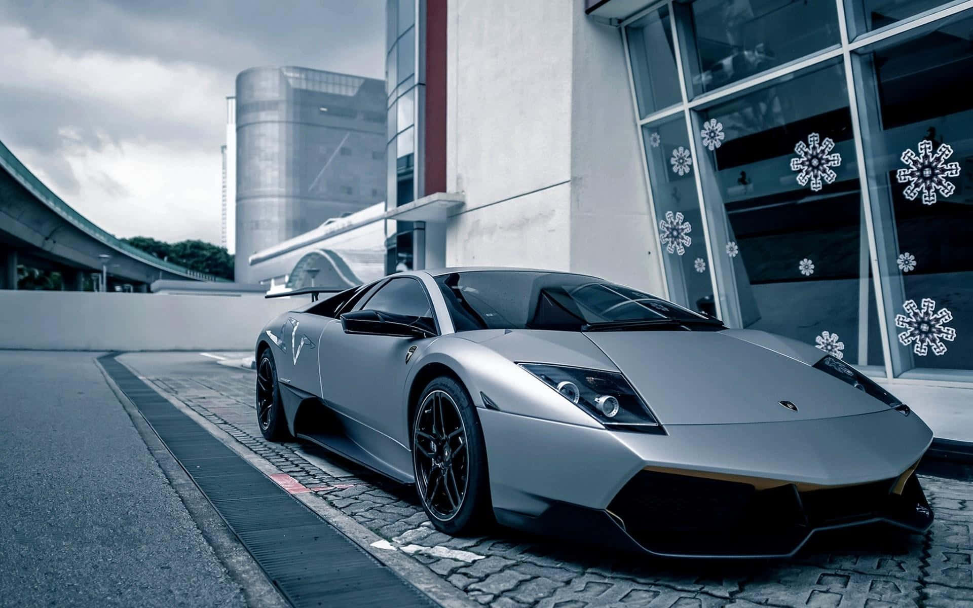 Lamborghini Murciélago 桌布 壁紙 — 一架時尚的銀色林寶堅尼Reventon停泊在鵝卵石路上，旁邊是一座現代建築，玻璃窗上有雪花貼紙，天空多雲。