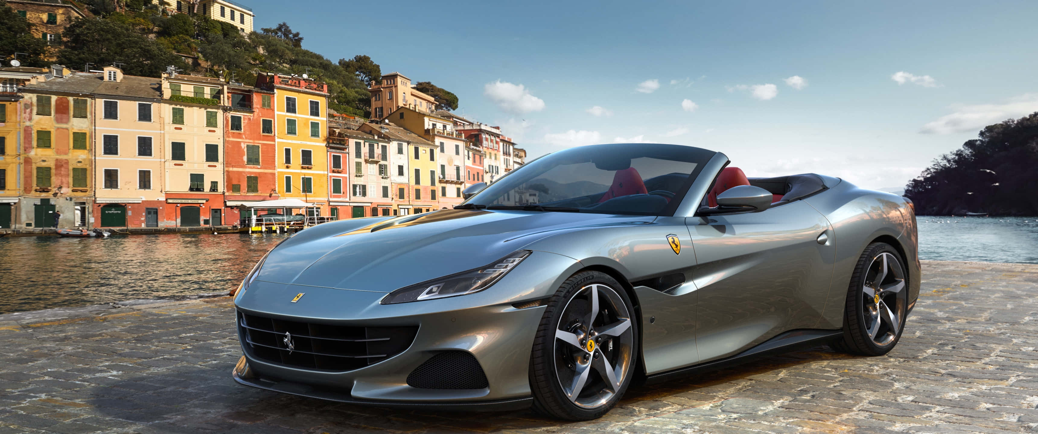 Ferrari Portofino 桌布 壁紙 — 一架時尚的銀色法拉利Portofino開篷跑車停泊在鵝卵石海濱，背景是色彩繽紛的意大利建築物和寧靜的水面。