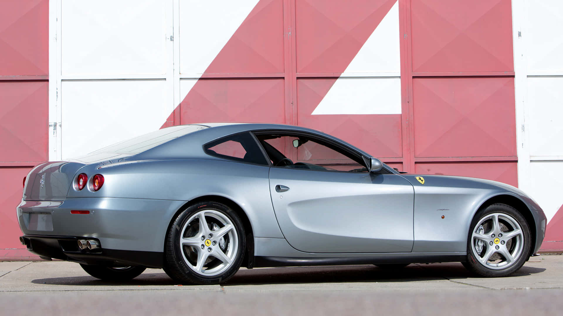 Ferrari 612 Scaglietti 桌布 壁紙 — 一架時尚的銀色法拉利跑車停放在大膽的紅白幾何牆前。