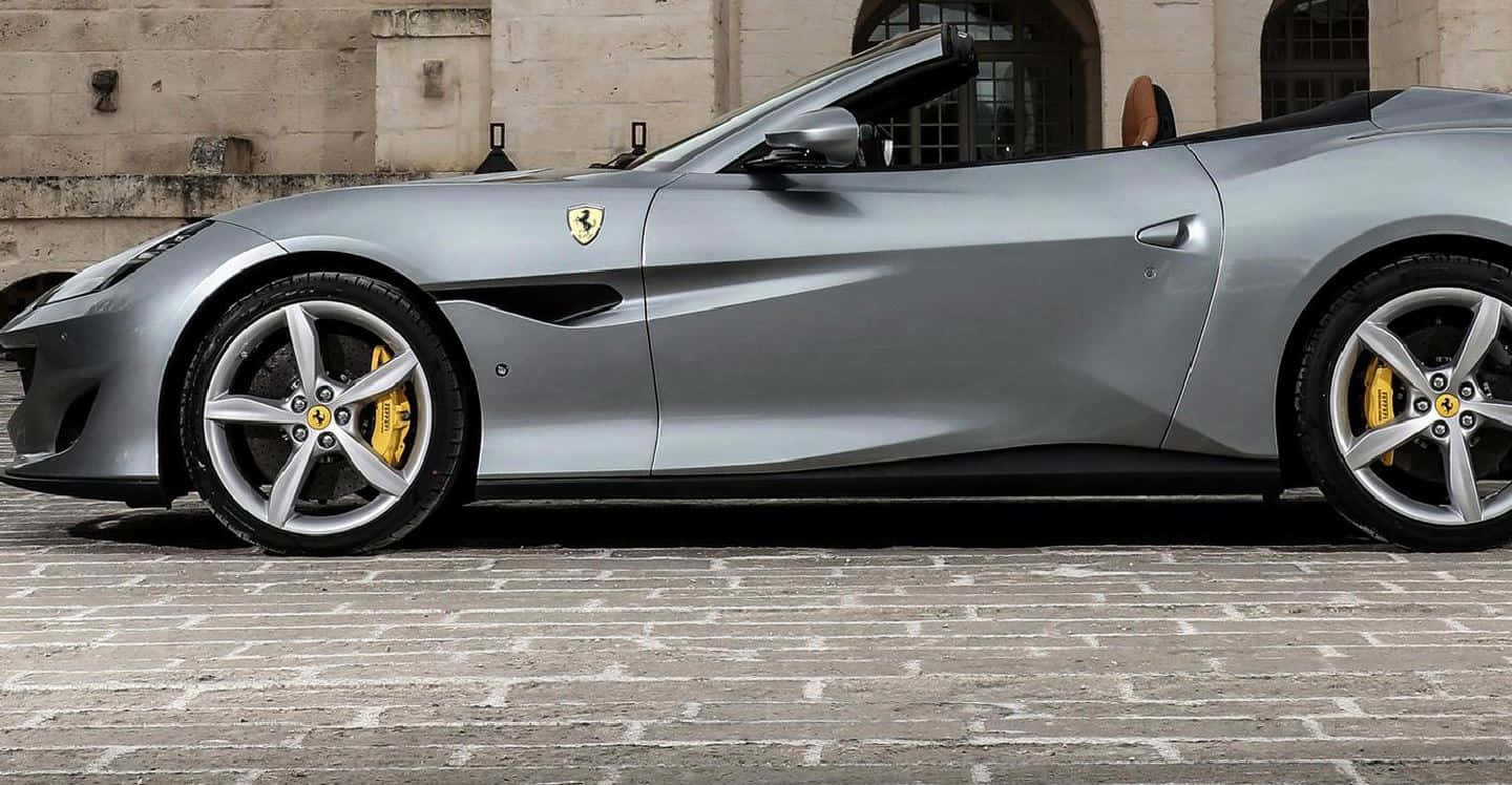 Ferrari Portofino 桌布 壁紙 — 一架時尚的銀色法拉利開篷車，帶有黃色制動卡鉗，停泊在石砌建築旁的鵝卵石街道上。