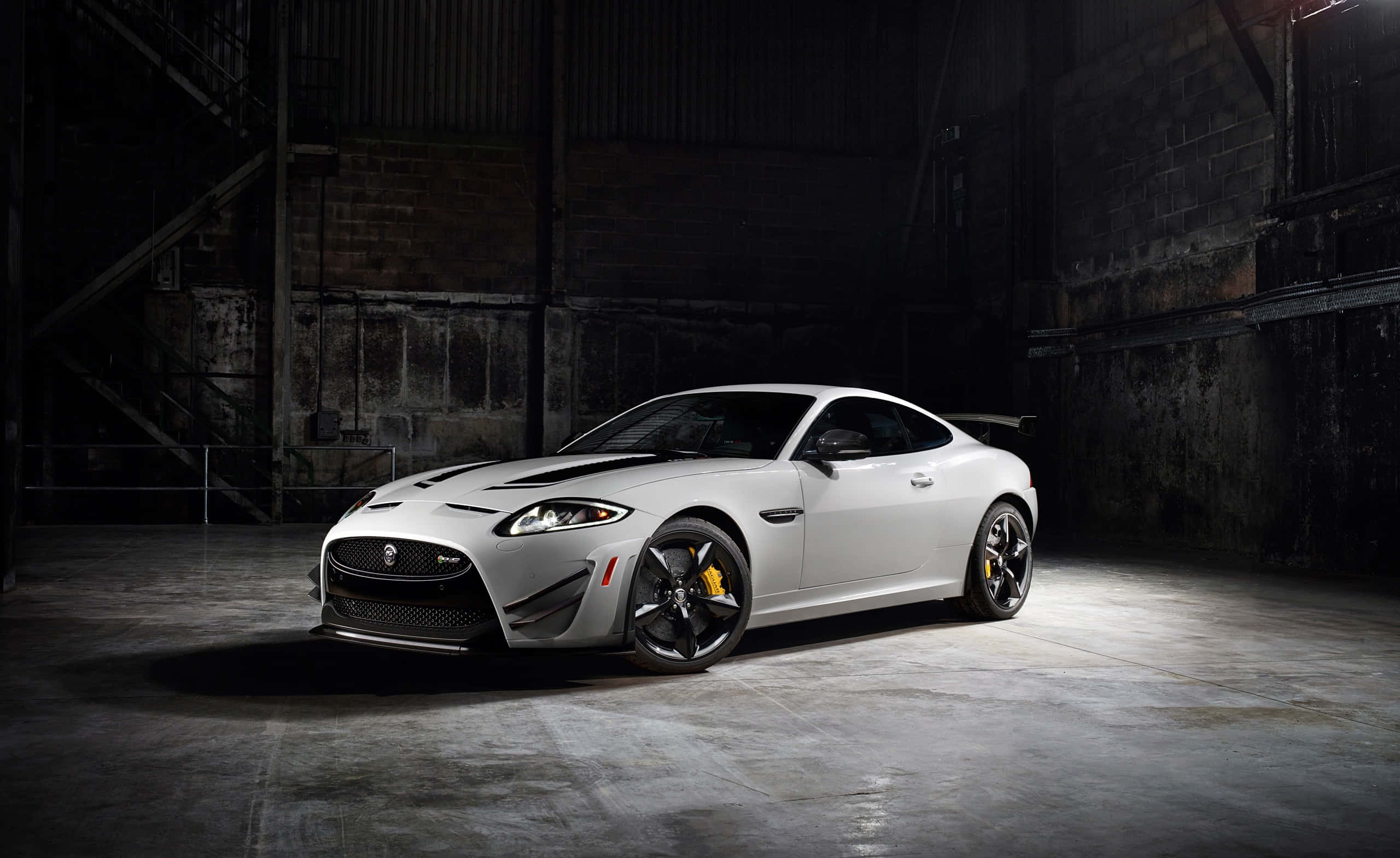 Jaguar Xkr 桌布 壁紙 — 一架時尚的銀色積架跑車，帶有黑色飾件和黃色制動卡鉗，在骯髒的工業倉庫中被戲劇性地照亮。