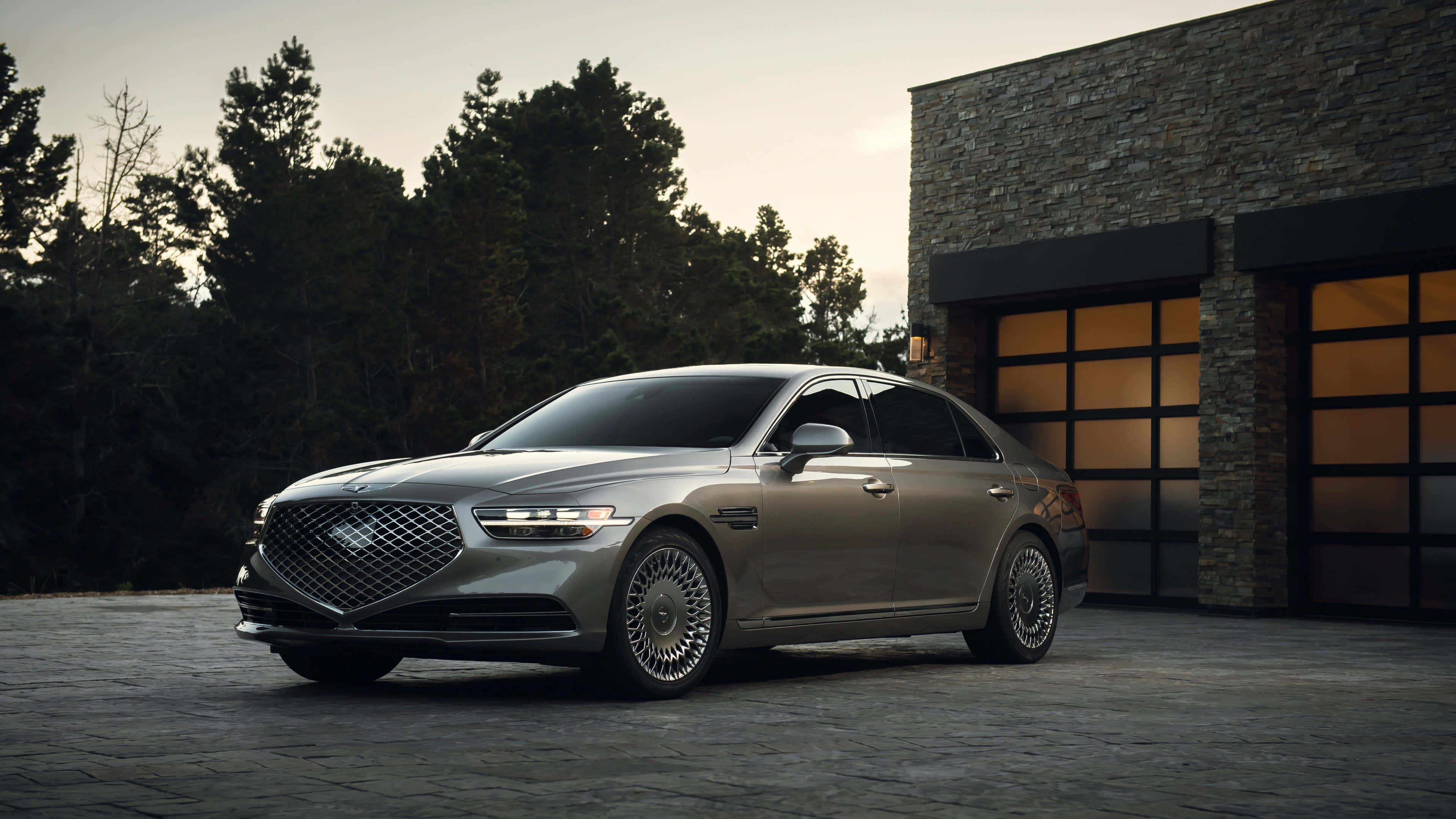 Genesis G90 桌布 壁紙 — 一架時尚的銀色豪華轎車停泊在石頭車道上，旁邊是一座帶有發光車庫門的現代石頭建築。