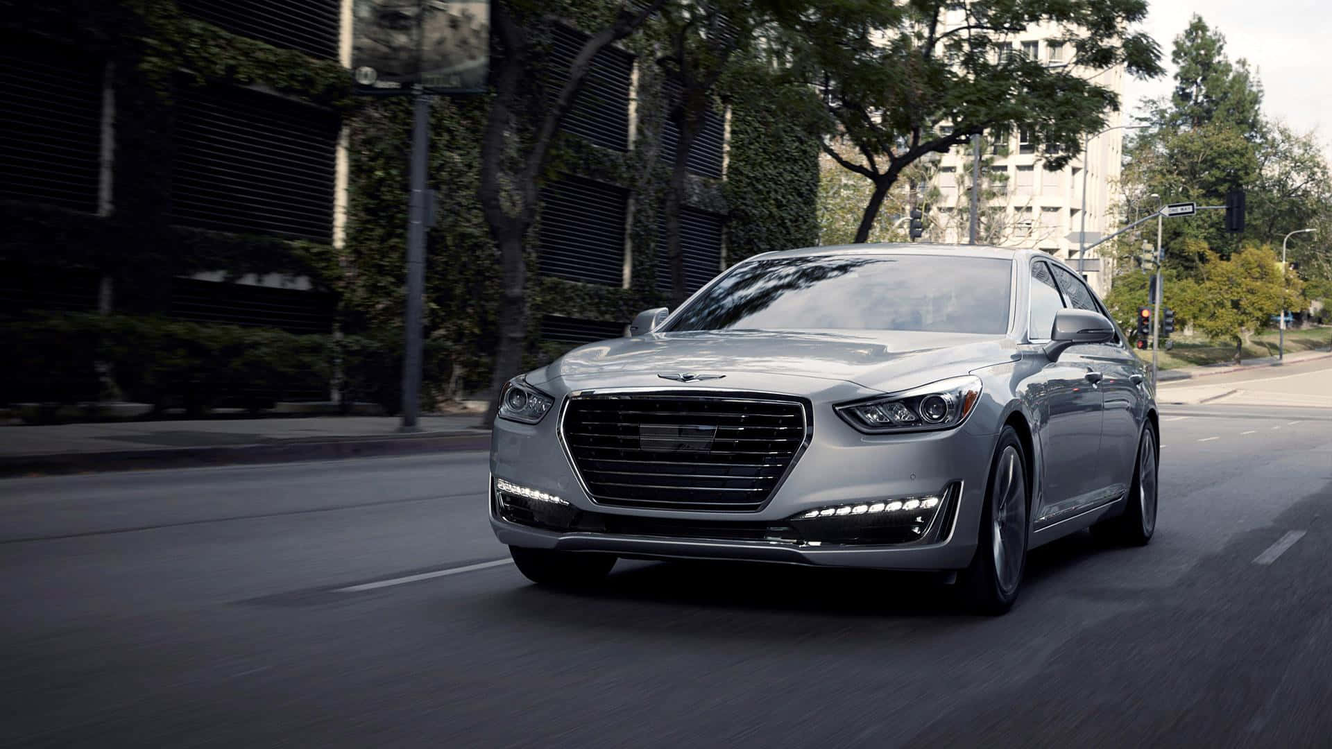 Genesis G90 桌布 壁紙 — 一架時尚的銀色豪華轎車駛過綠樹成蔭的市區街道，背景是現代建築。